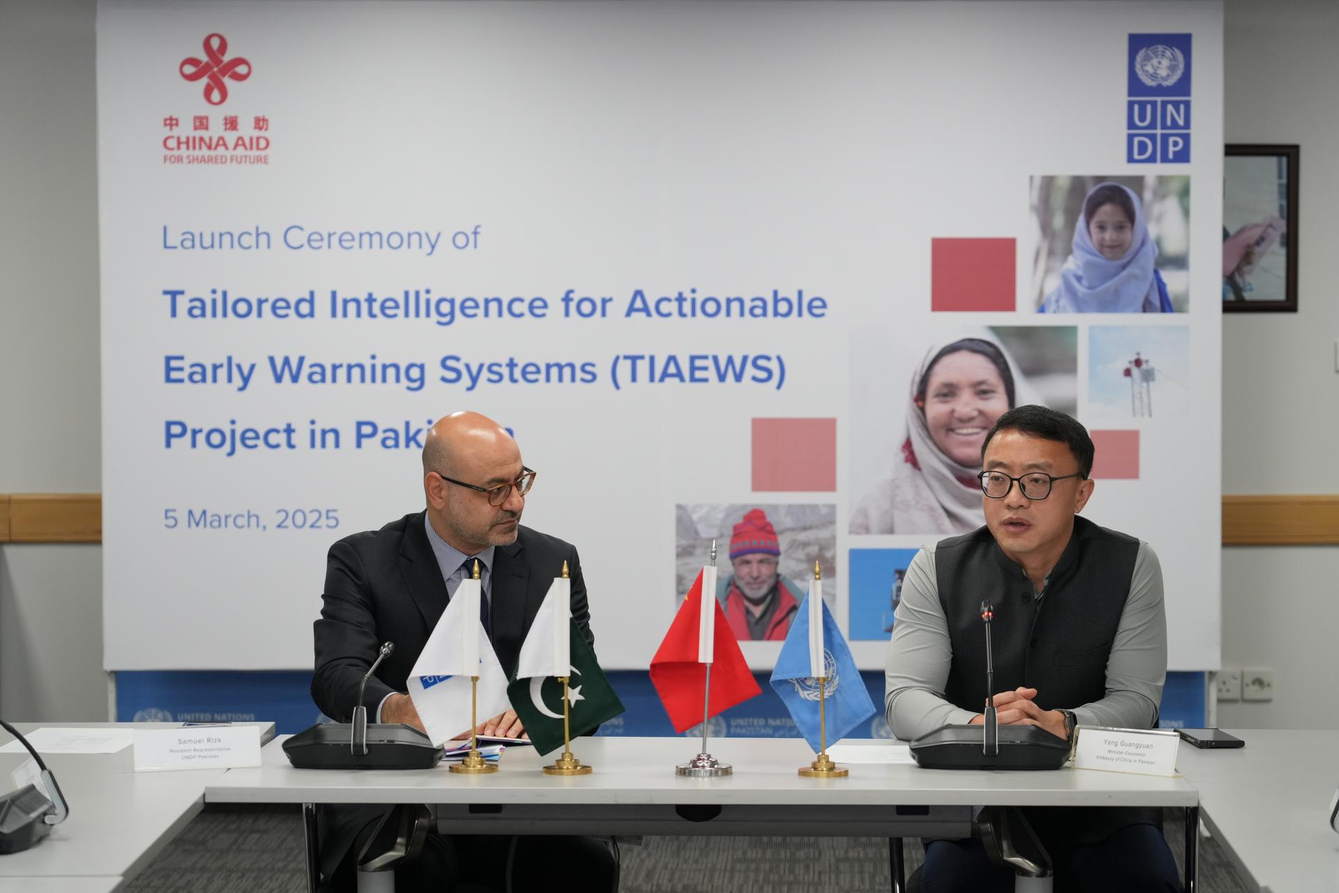 Two men seated at a table during the launch of a project, with flags and a backdrop featuring various individuals.