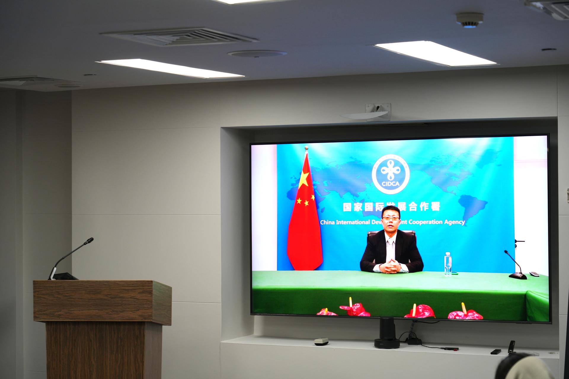 A man sits at a table in front of a large screen displaying a flag and logo.
