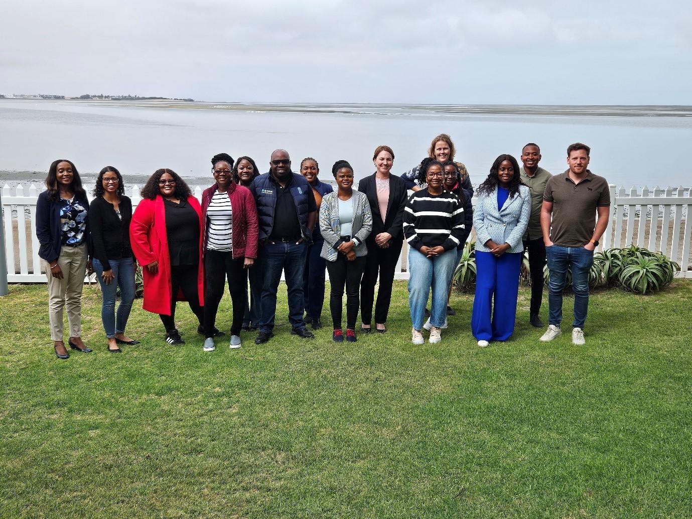 Group of diverse individuals posed together outdoors by a body of water, smiling.