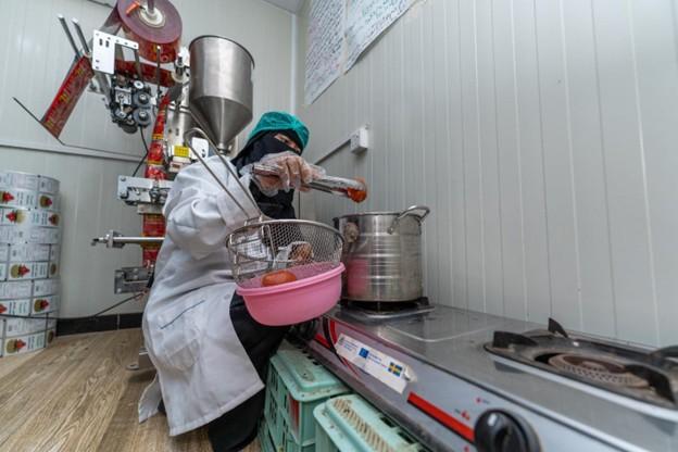 A person in a lab coat and mask prepares food in a kitchen, surrounded by equipment.
