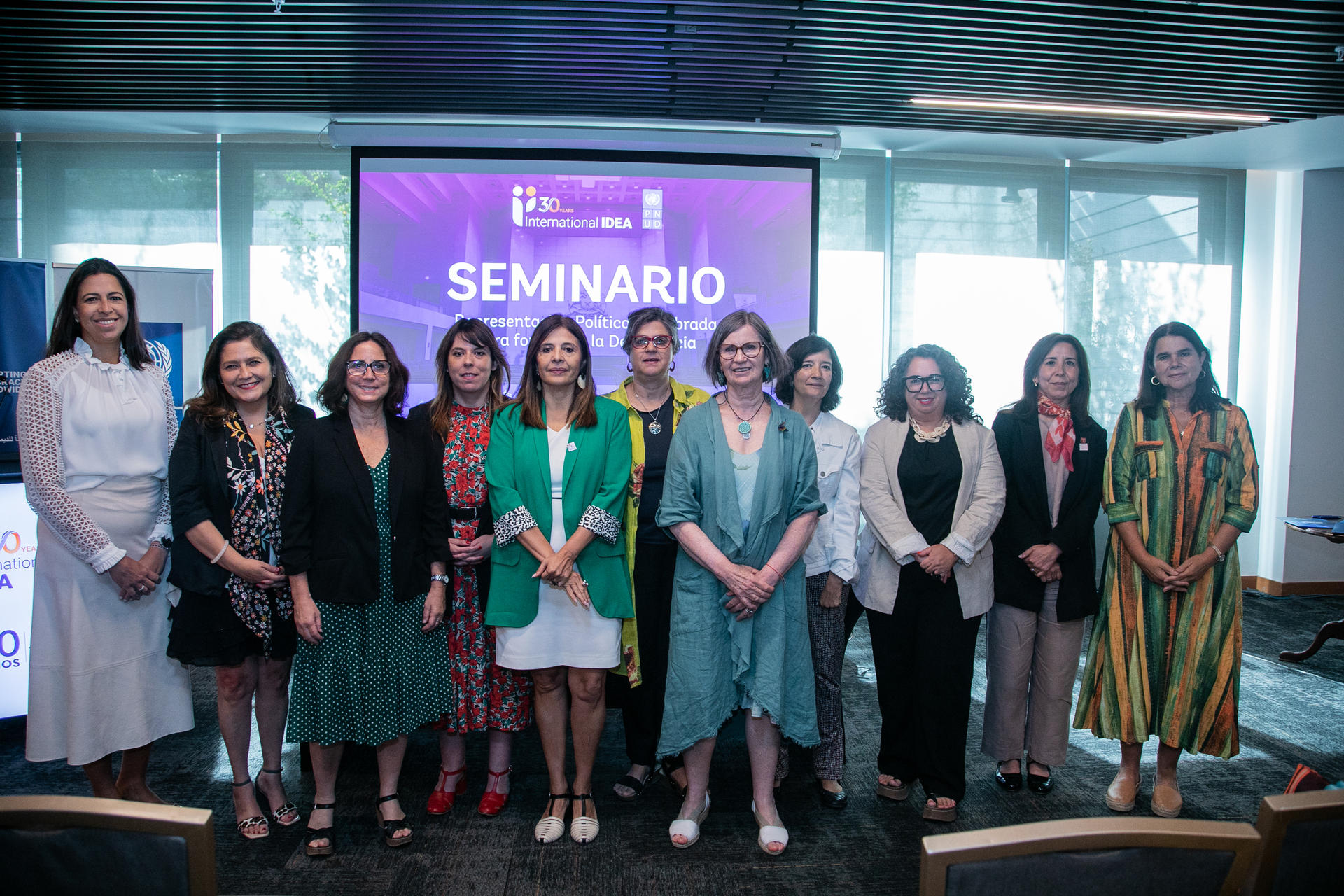 A group of diverse women stands together in a well-lit seminar room, smiling.