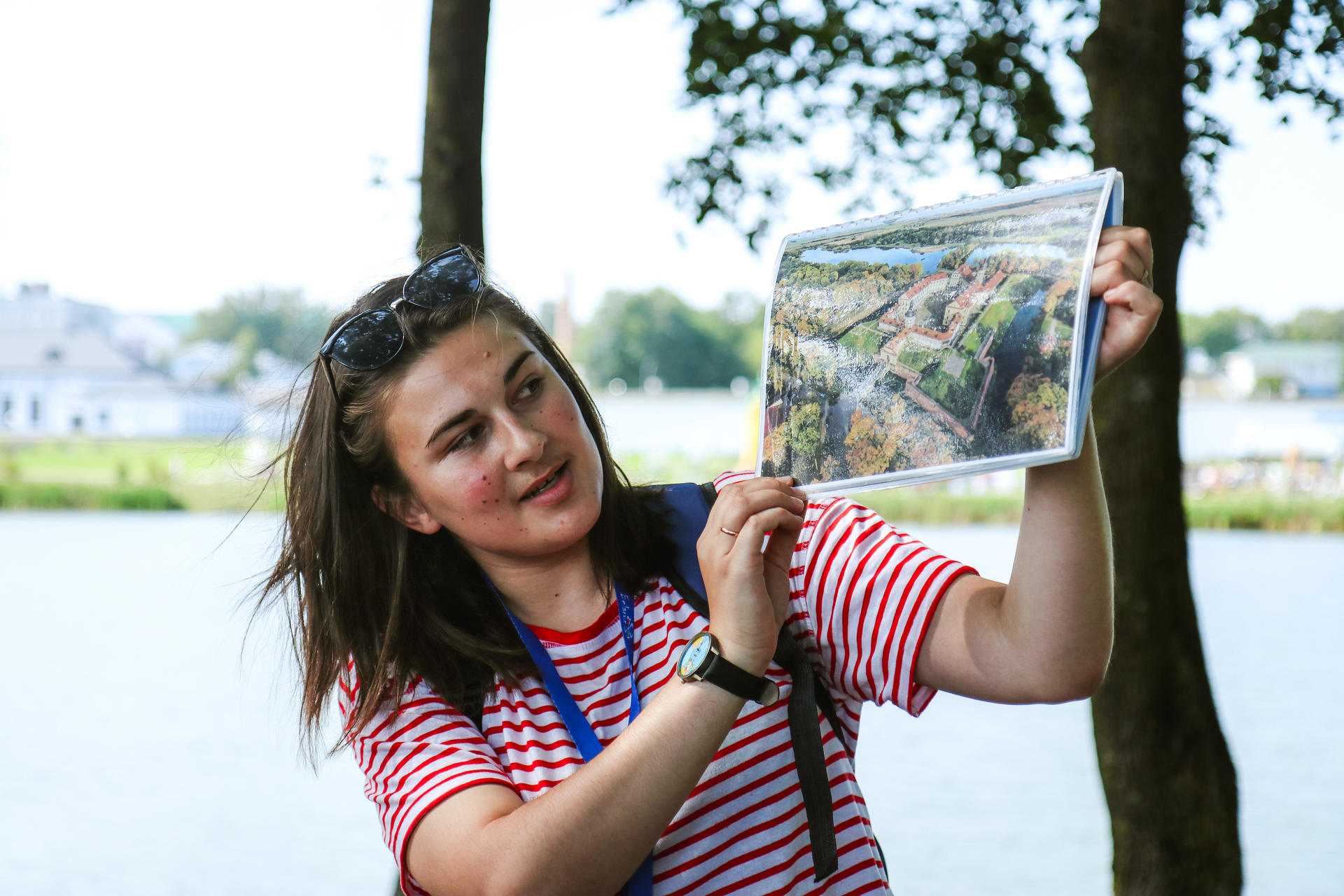 Young woman in striped shirt holds a landscape photo by a lake, surrounded by trees.