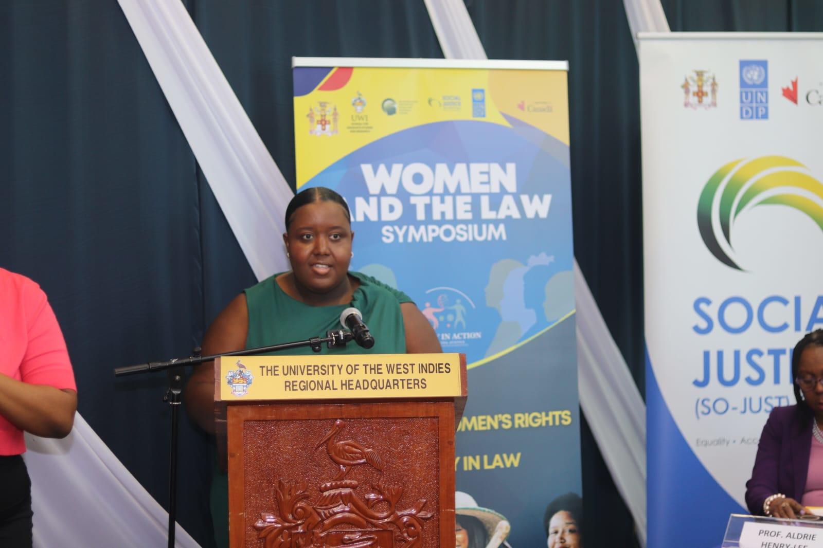 Woman speaking at a podium during the Women and the Law Symposium, with banners in the background.