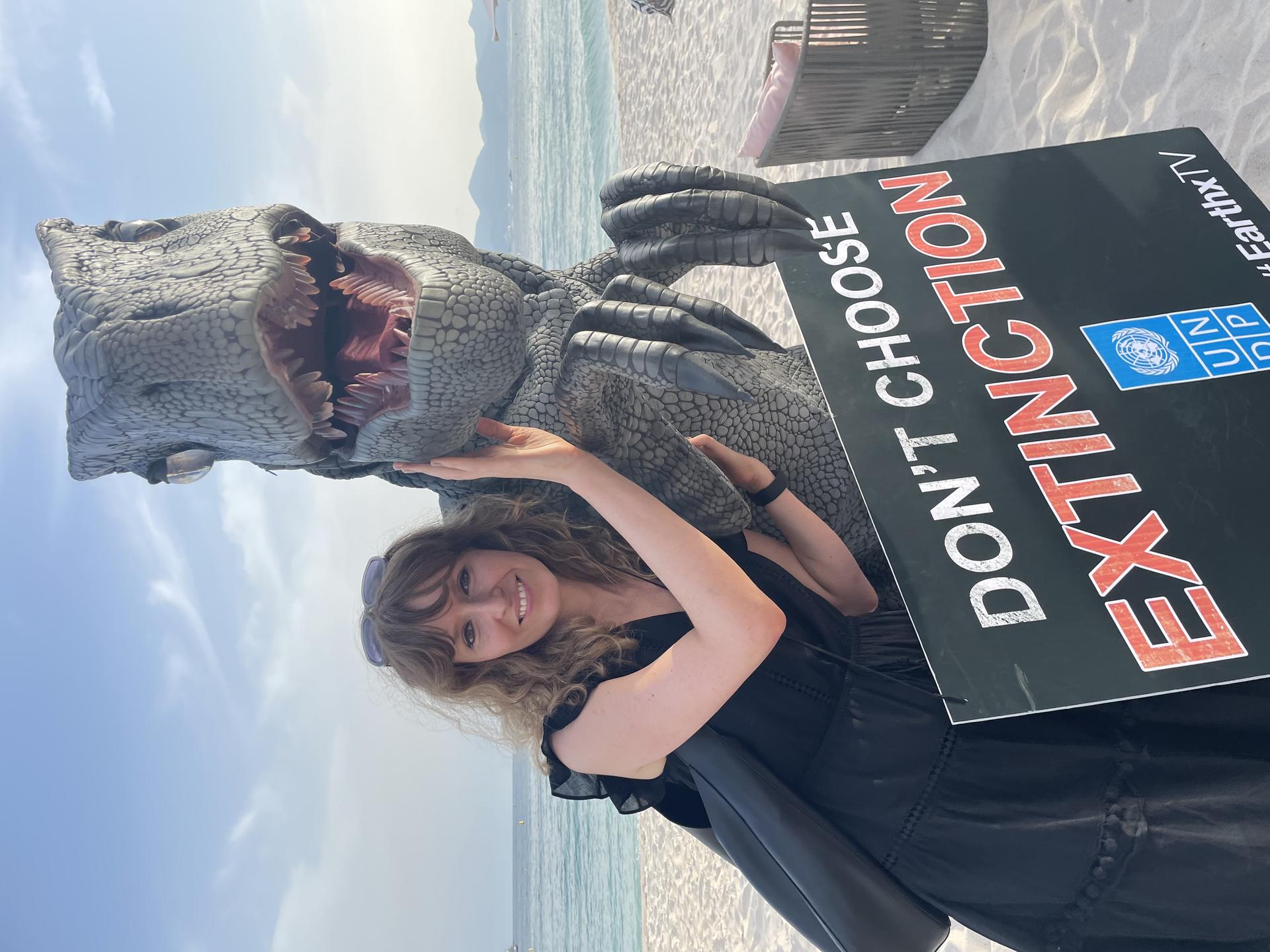 A woman poses with a dinosaur costume and a protest sign on a beach.
