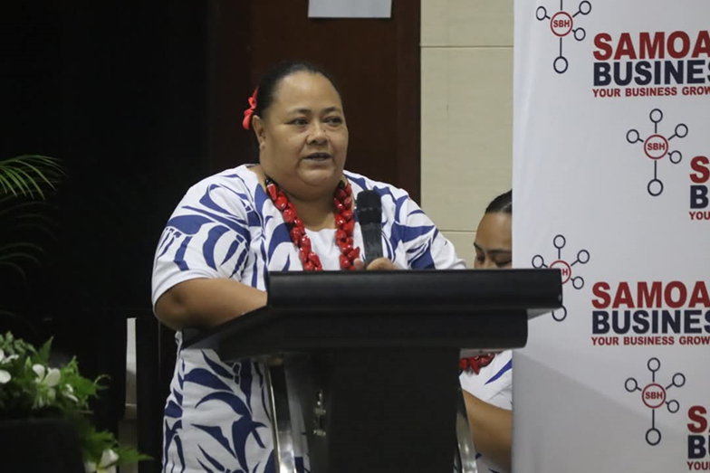 Woman speaking at a podium, wearing a blue and white patterned dress and red necklace.
