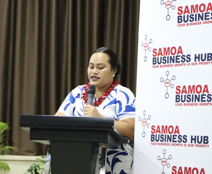 A woman in a blue and white patterned dress speaks at a podium beside a Samoa Business Hub banner.