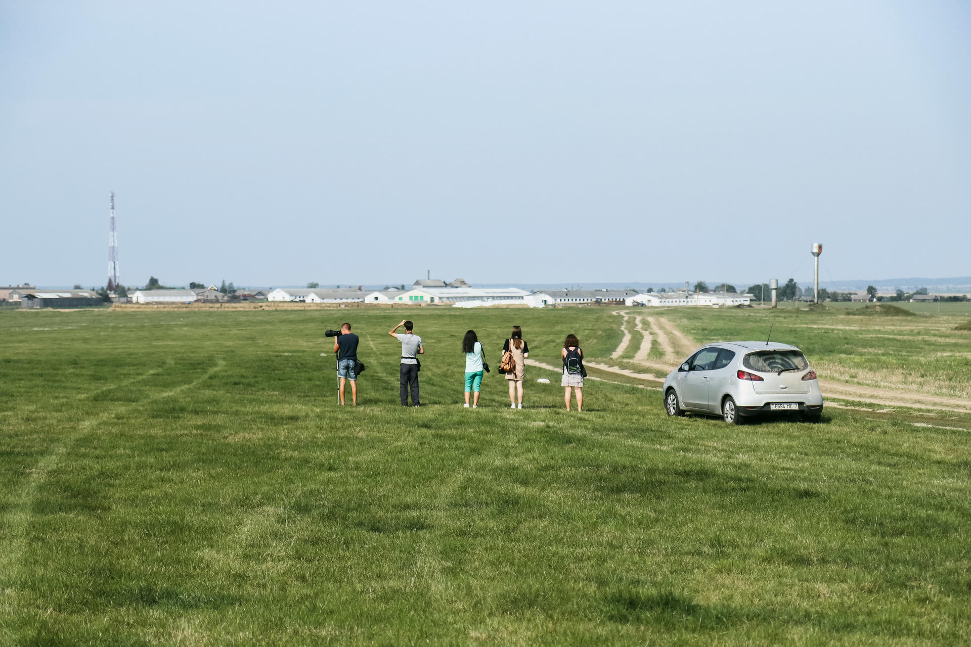 A group of people stands in a grassy field, facing a distant landscape with a parked car nearby.