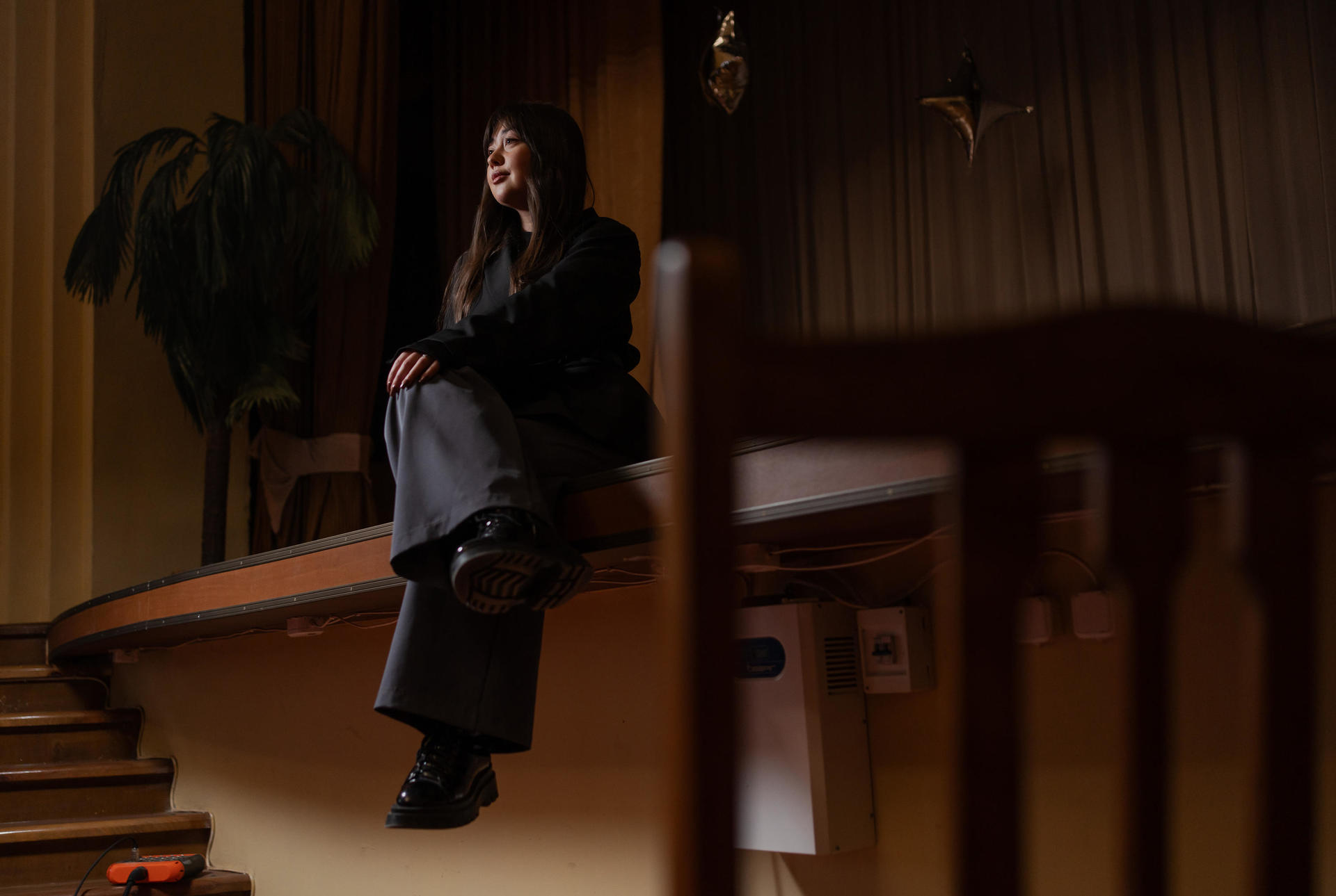 A young woman sits on a stage, gazing pensively, with soft lighting and a plant in the background.
