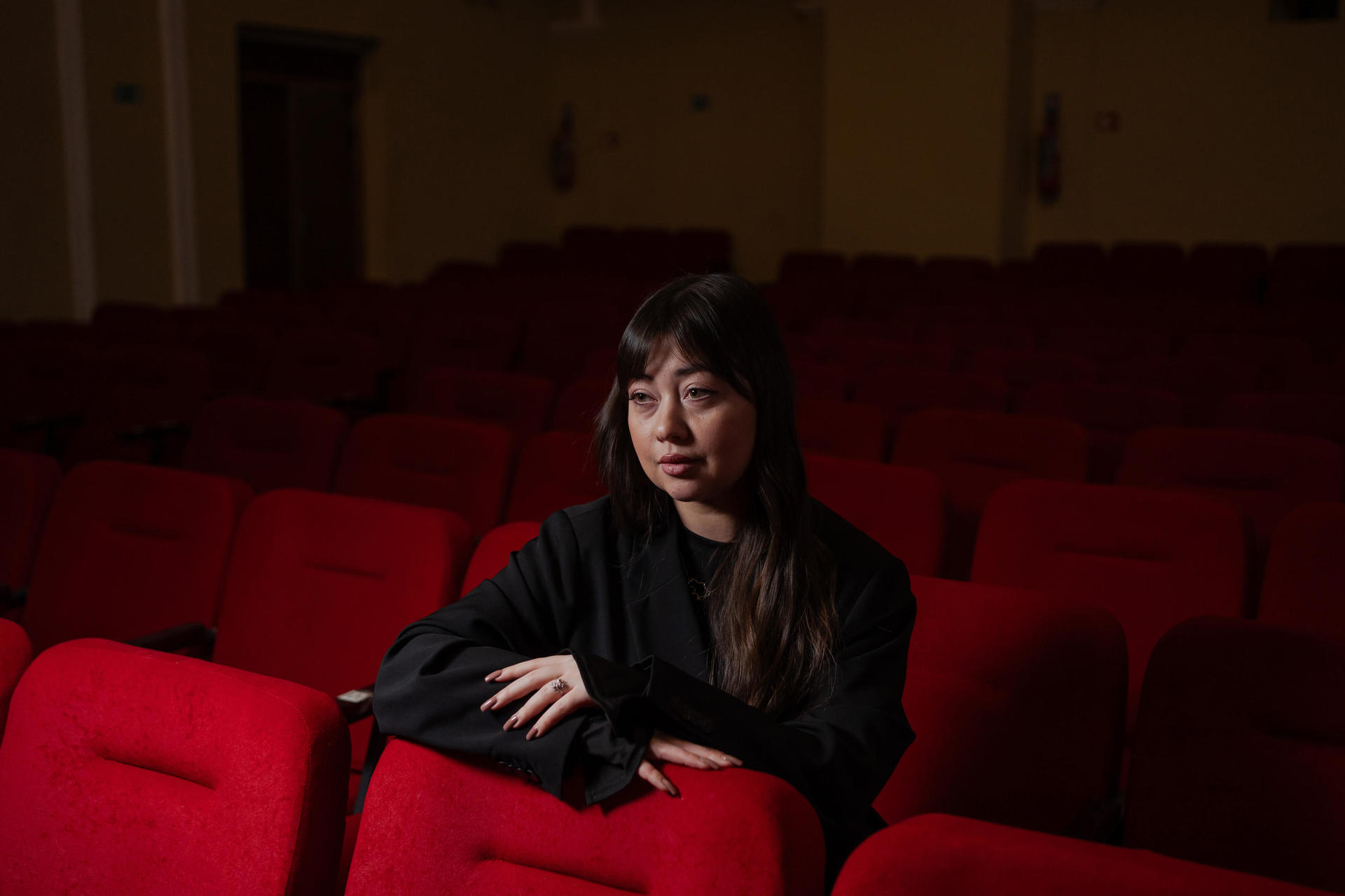 A woman sits thoughtfully in an empty theater, surrounded by red seats.