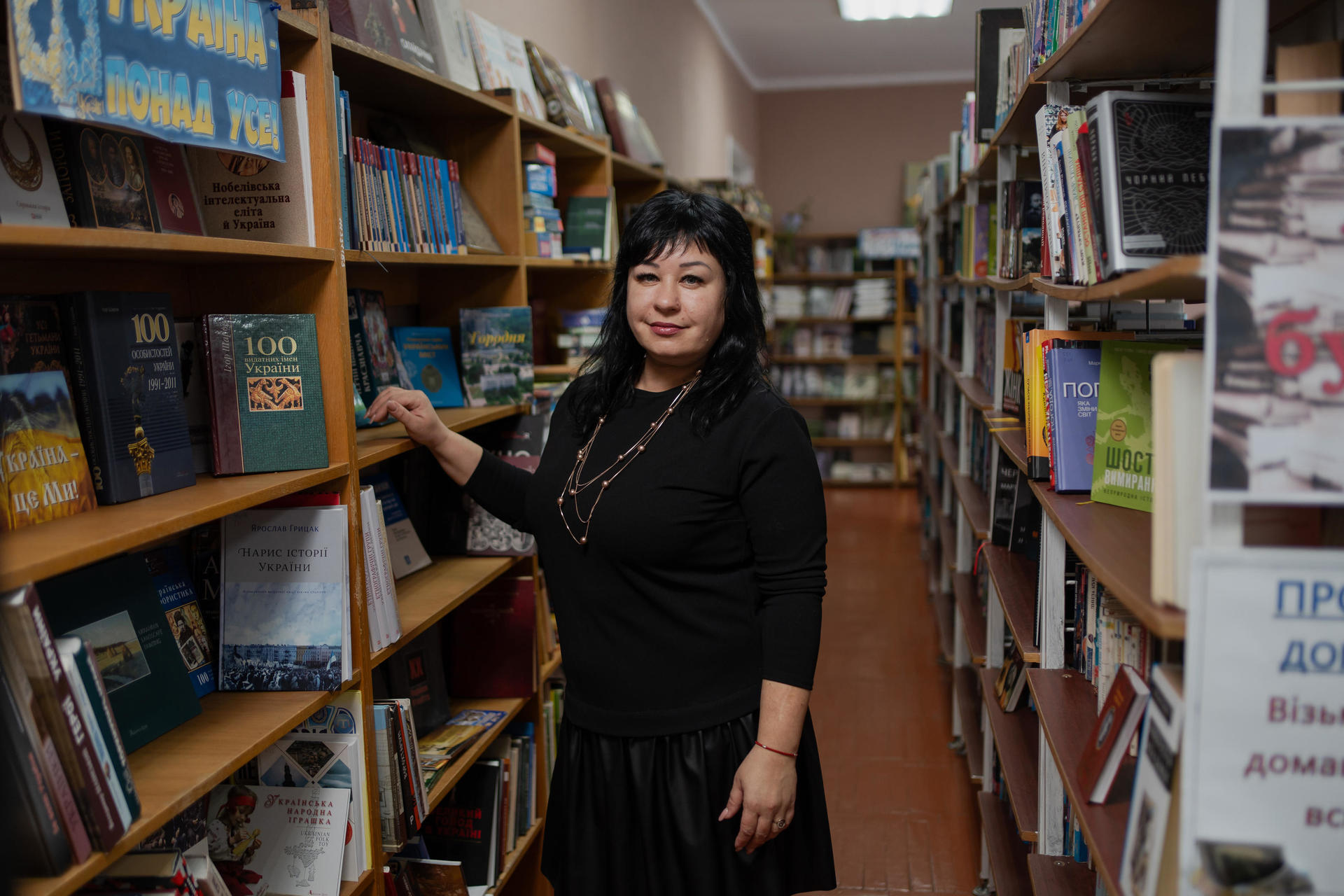 Woman standing beside a bookshelf in a library, smiling and holding a book.