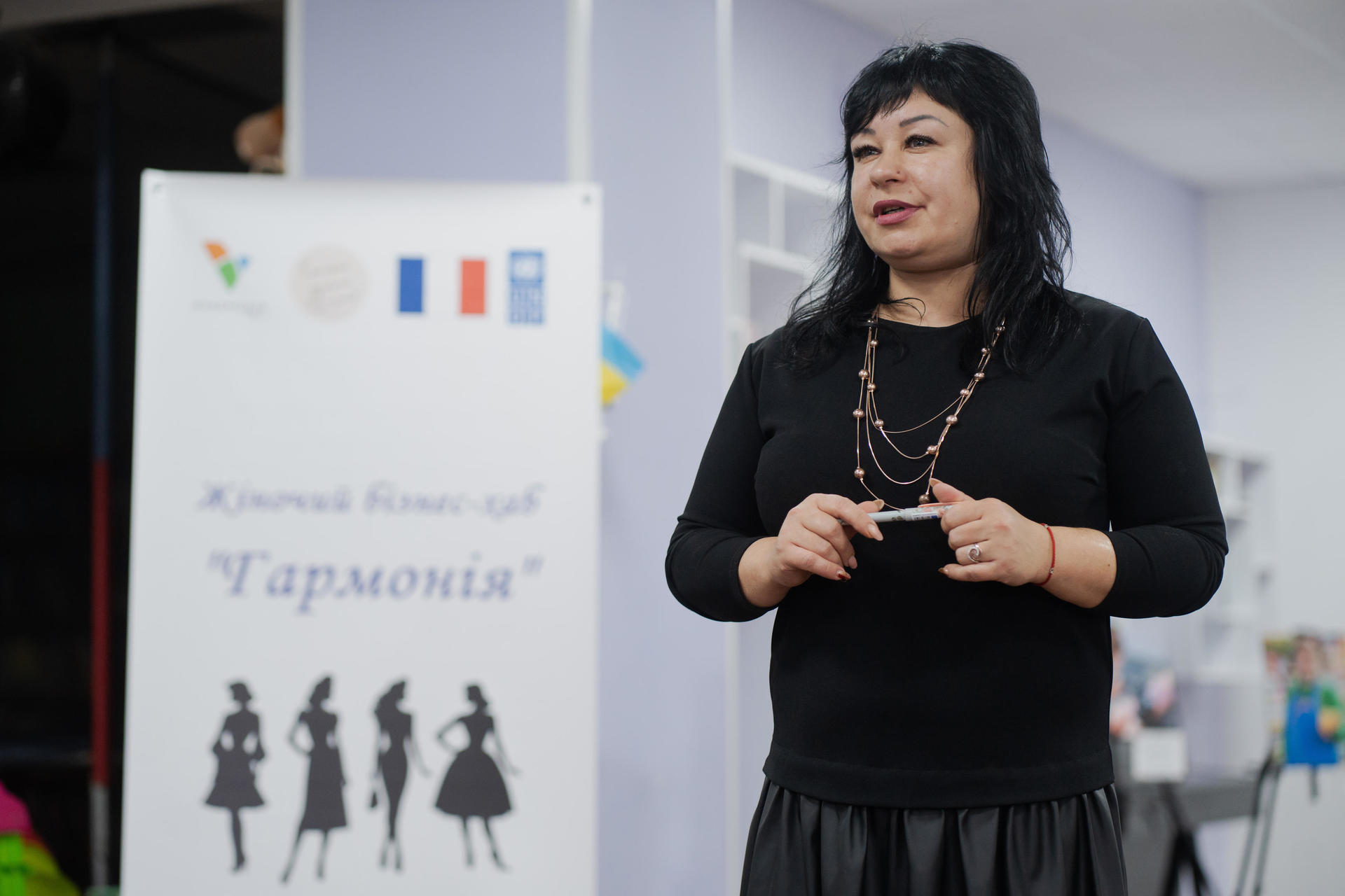 A woman with dark hair wearing a black top speaks in a colorful room with a banner.