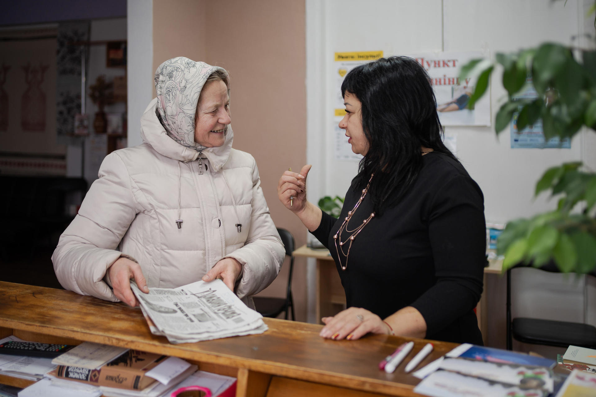 Two women engage in conversation at a counter, one holding a newspaper.