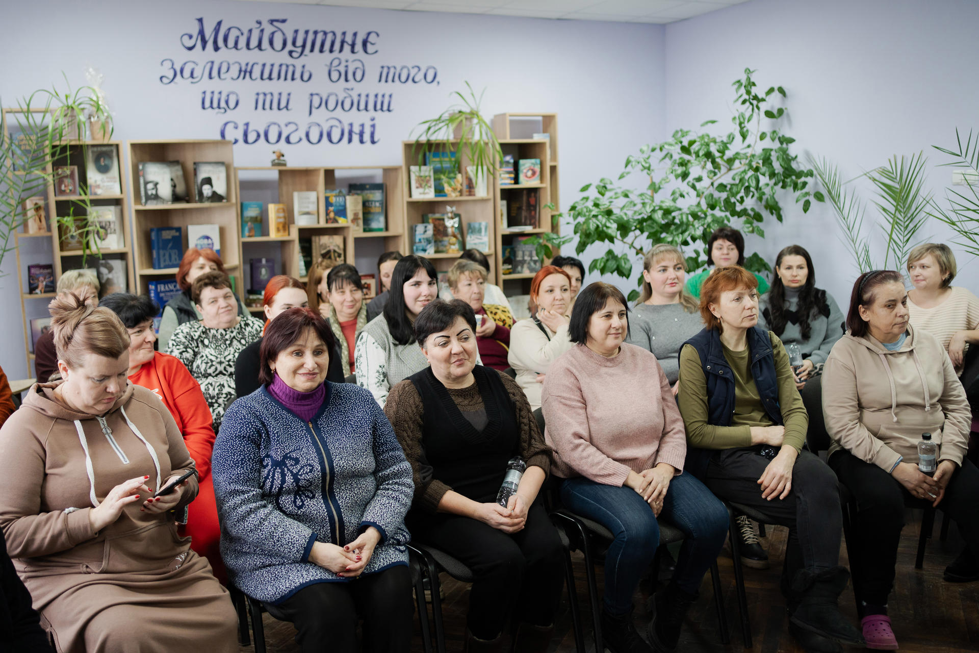 A group of women seated in a room with bookshelves and greenery, looking engaged.