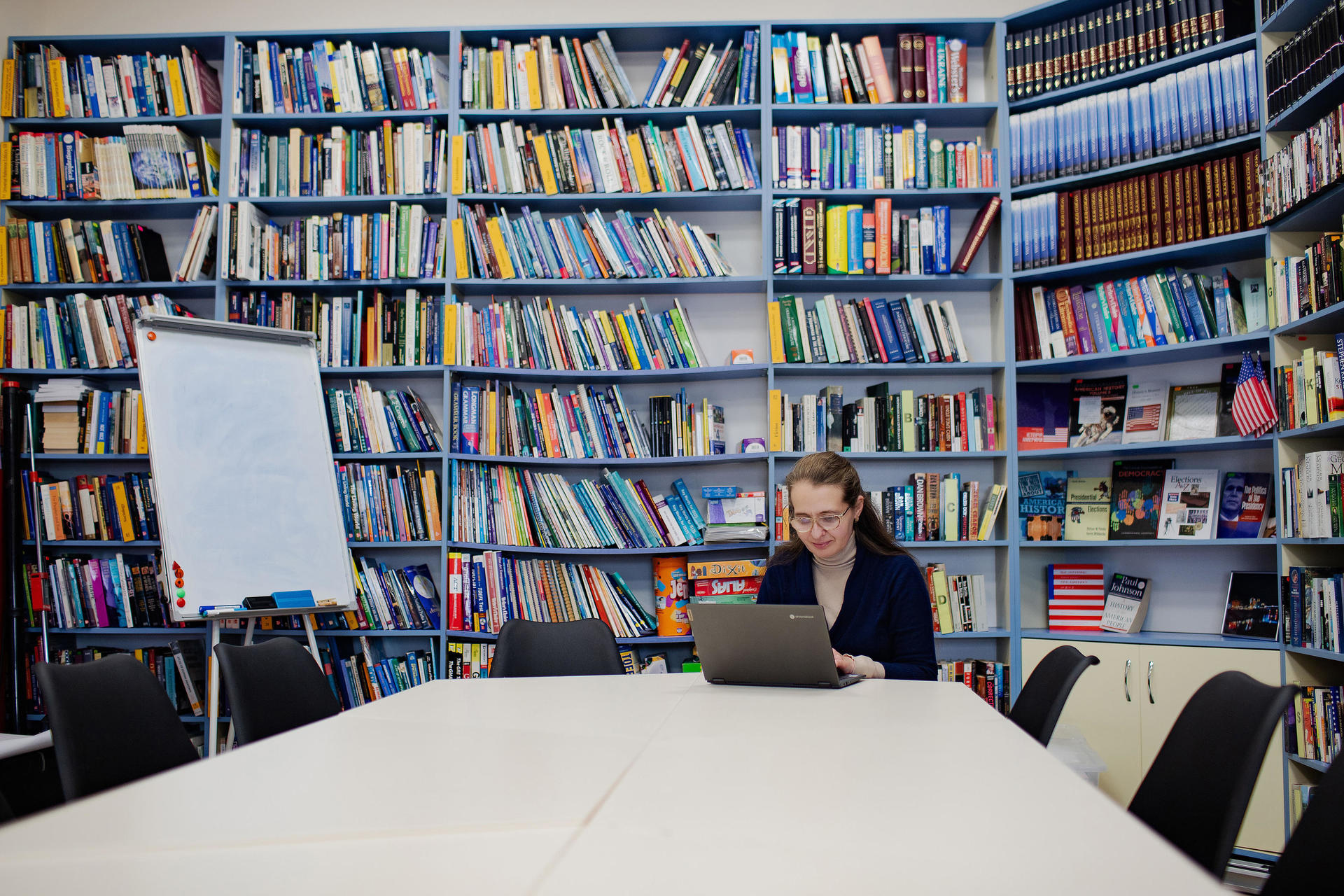 A woman works on a laptop at a table in a book-filled library. Shelves are colorful and well-stocked.