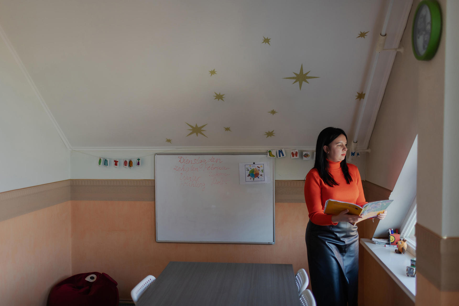 A woman in an orange sweater reads a book by a window in a cozy, bright classroom.