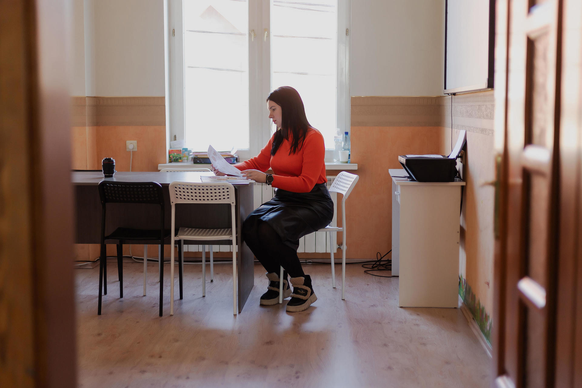 A woman in a red shirt sits at a table reading documents in a bright room.