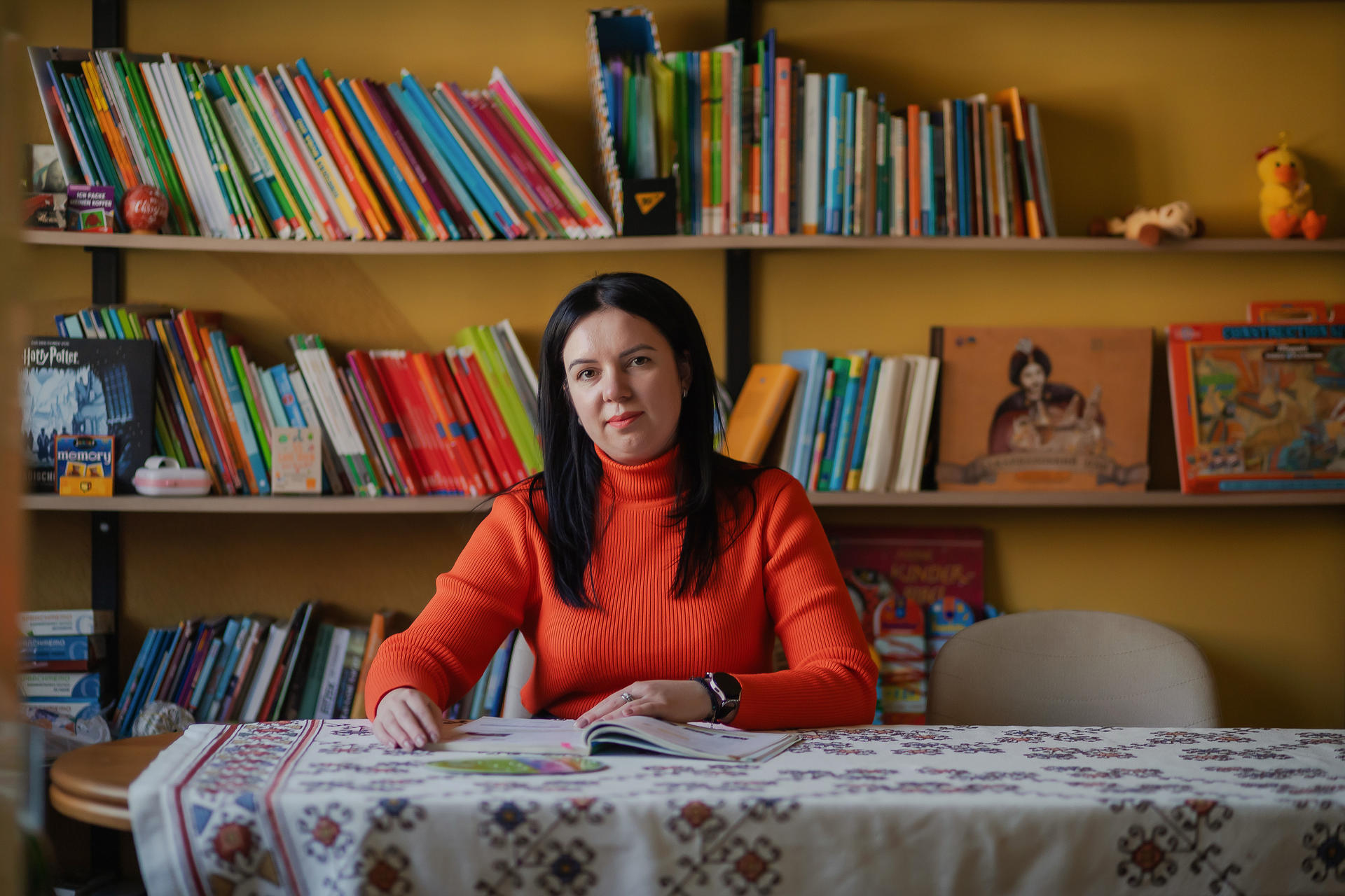 A woman in an orange sweater sits at a table in front of colorful bookshelves.