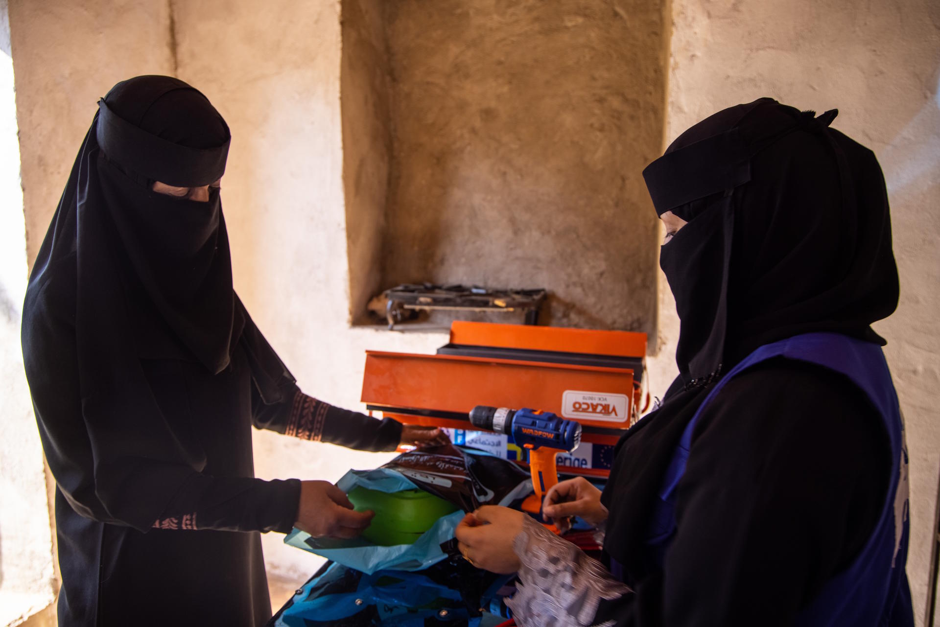 Two women in black abayas are discussing items at a table in a well-lit, rustic space.