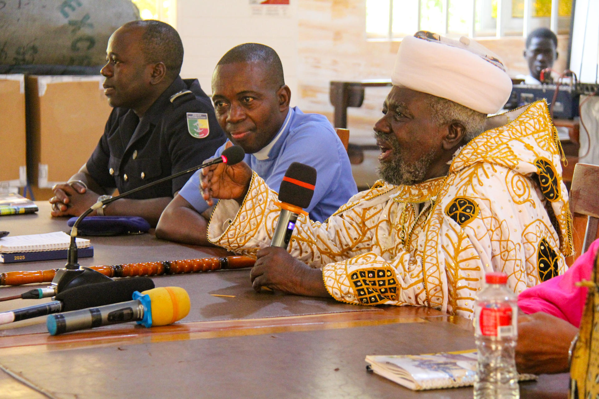A group of men seated at a table during a discussion; one man in traditional attire speaks.
