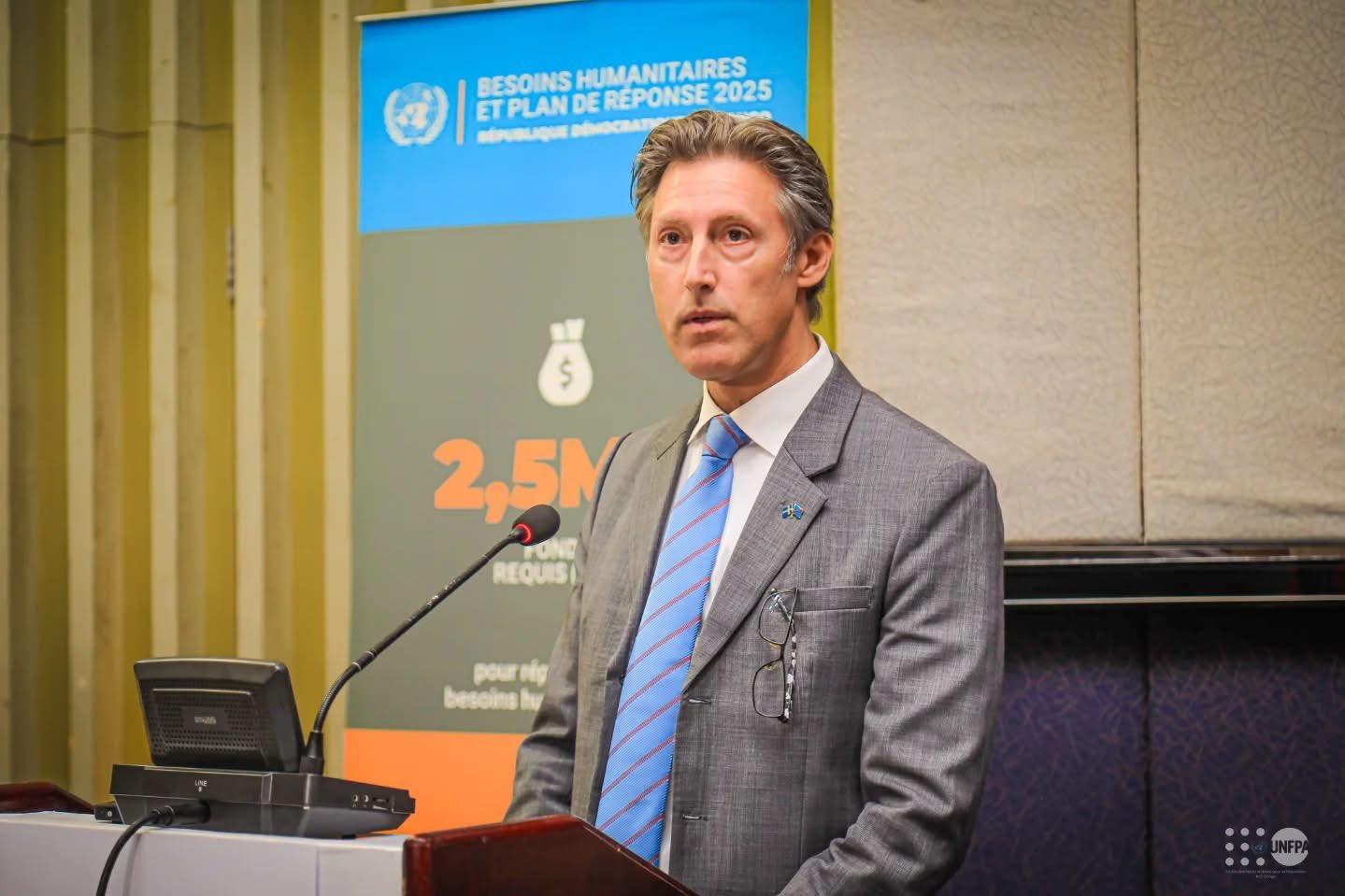 A man in a suit stands at a podium, speaking at a conference with a banner in the background.