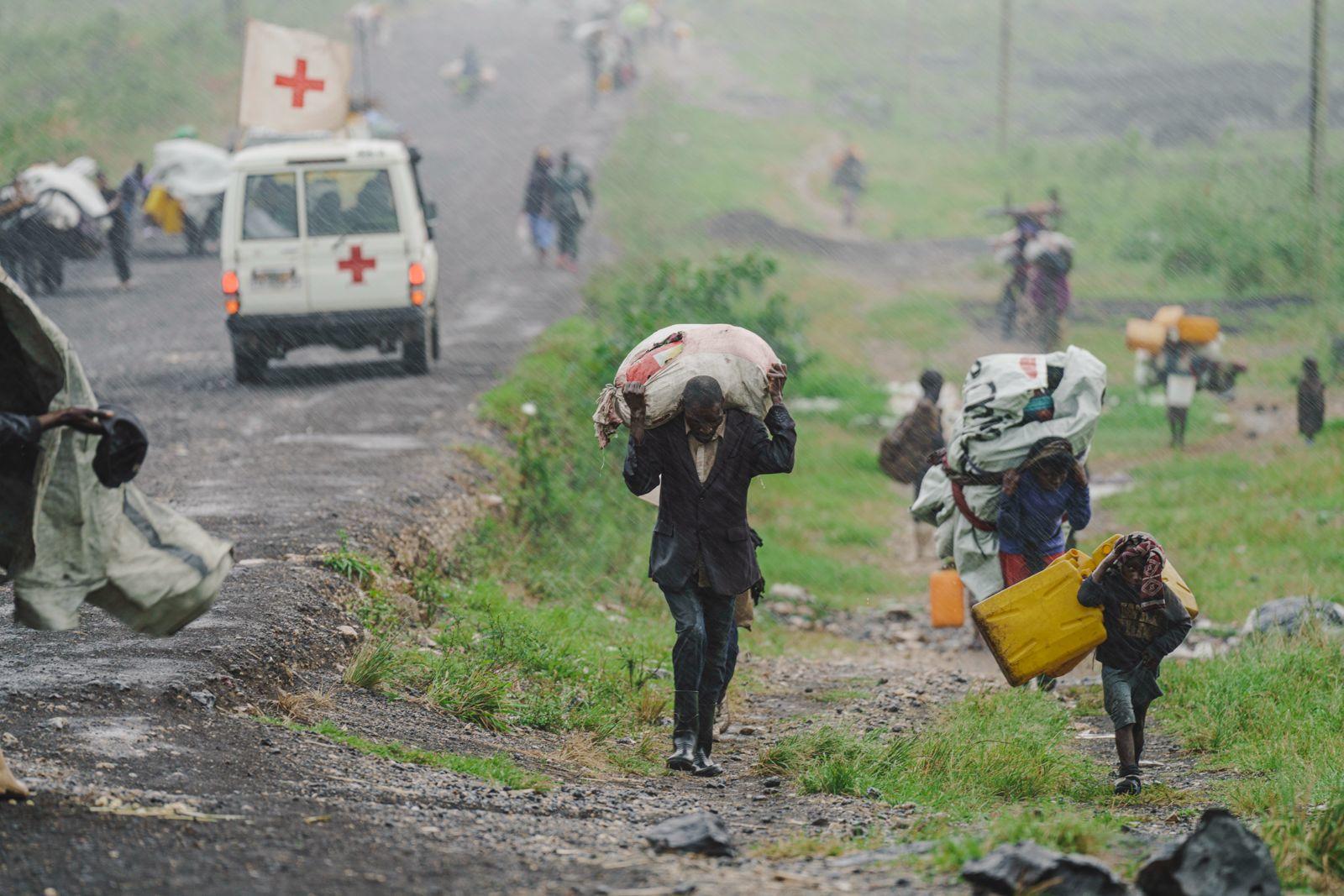 People walking along a muddy road carrying heavy loads, with a Red Cross vehicle nearby.