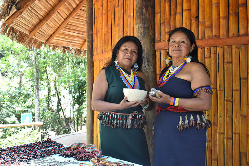 Two women in traditional attire stand in front of a bamboo structure, holding a bowl.