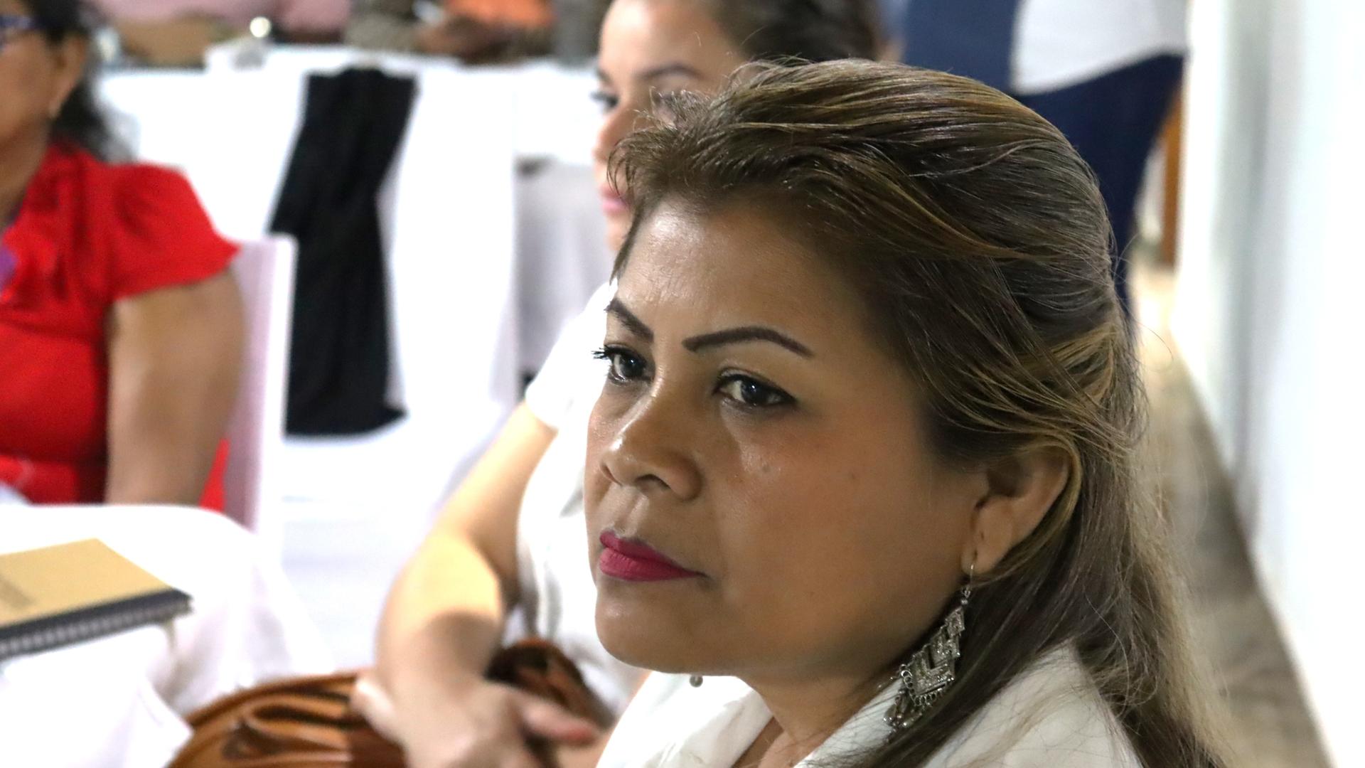 AUna mujer de cabello oscuro y aretes mira pensativamente hacia un lado en una reunión.