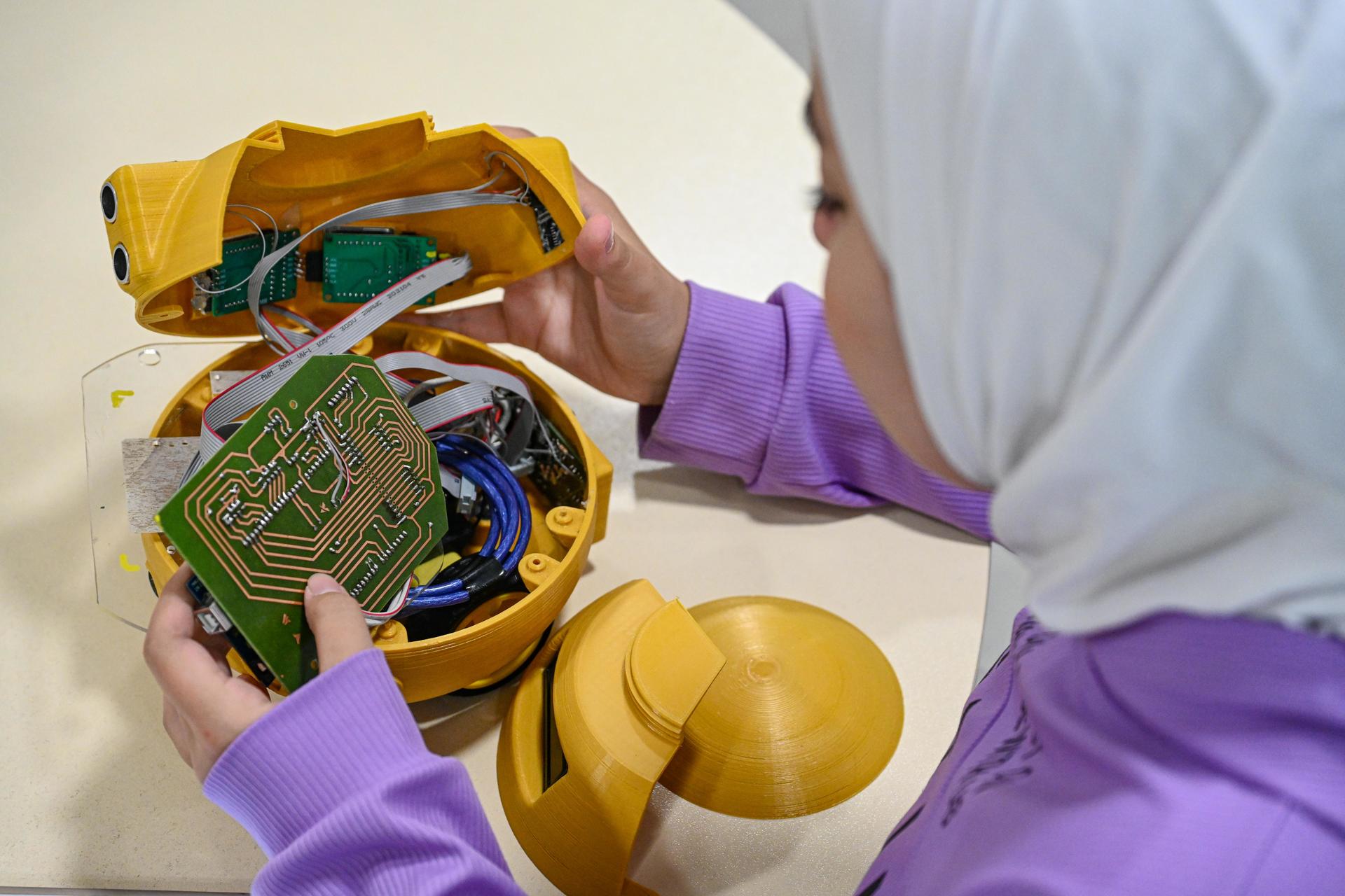 A girl in a hijab examines the inside of a yellow robotic toy with wires and a circuit board.