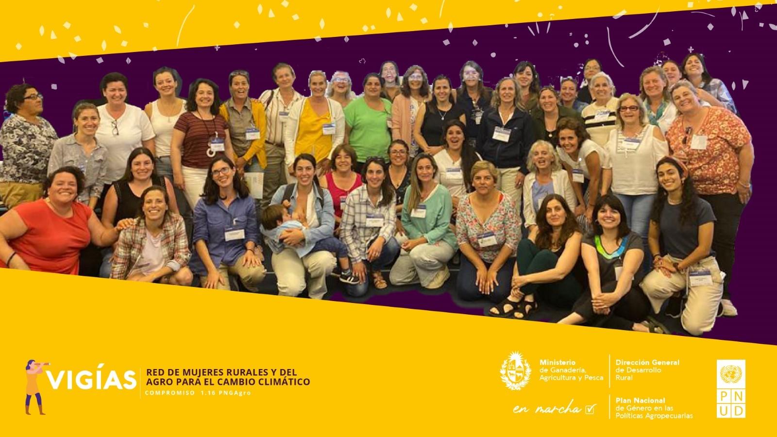Fotografía grupal de mujeres participantes en la red 'VIGÍAS: Red de Mujeres Rurales y del Agro para el Cambio Climático'. Aparecen sonriendo y en actitud de camaradería, algunas de pie y otras sentadas, con una madre sosteniendo a su hijo en brazos. La imagen tiene un diseño gráfico con fondo amarillo y morado, con elementos festivos como confeti. Se incluyen los logotipos del Ministerio de Ganadería, Agricultura y Pesca de Uruguay, la Dirección General de Desarrollo Rural, el Plan Nacional de Género en la