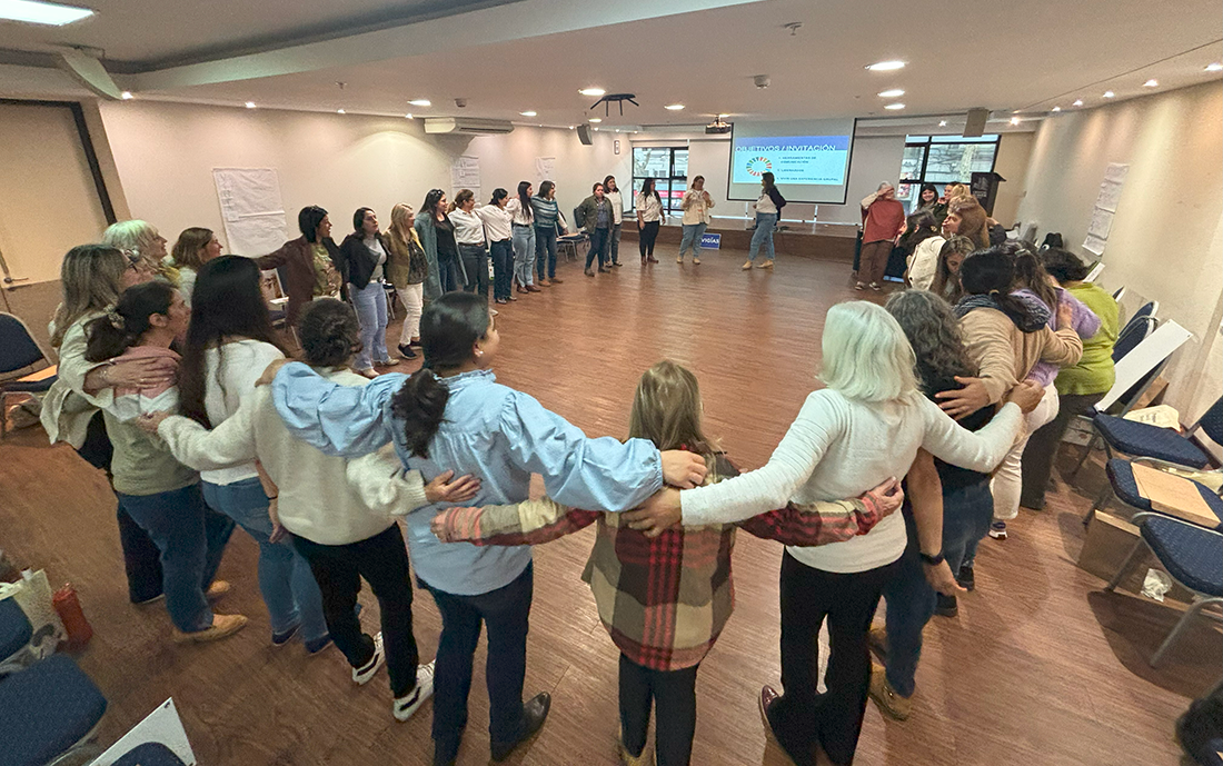 Fotografía de un grupo de mujeres formando un gran círculo en un salón de reuniones, abrazándose por los hombros en un gesto de unión y compañerismo. En el fondo, una pantalla proyecta una diapositiva con el título 'OBJETIVOS E INVITACIÓN'. La escena transmite un ambiente de colaboración y fortalecimiento comunitario, en lo que parece ser un encuentro de la red 'VIGÍAS: Red de Mujeres Rurales y del Agro para el Cambio Climático