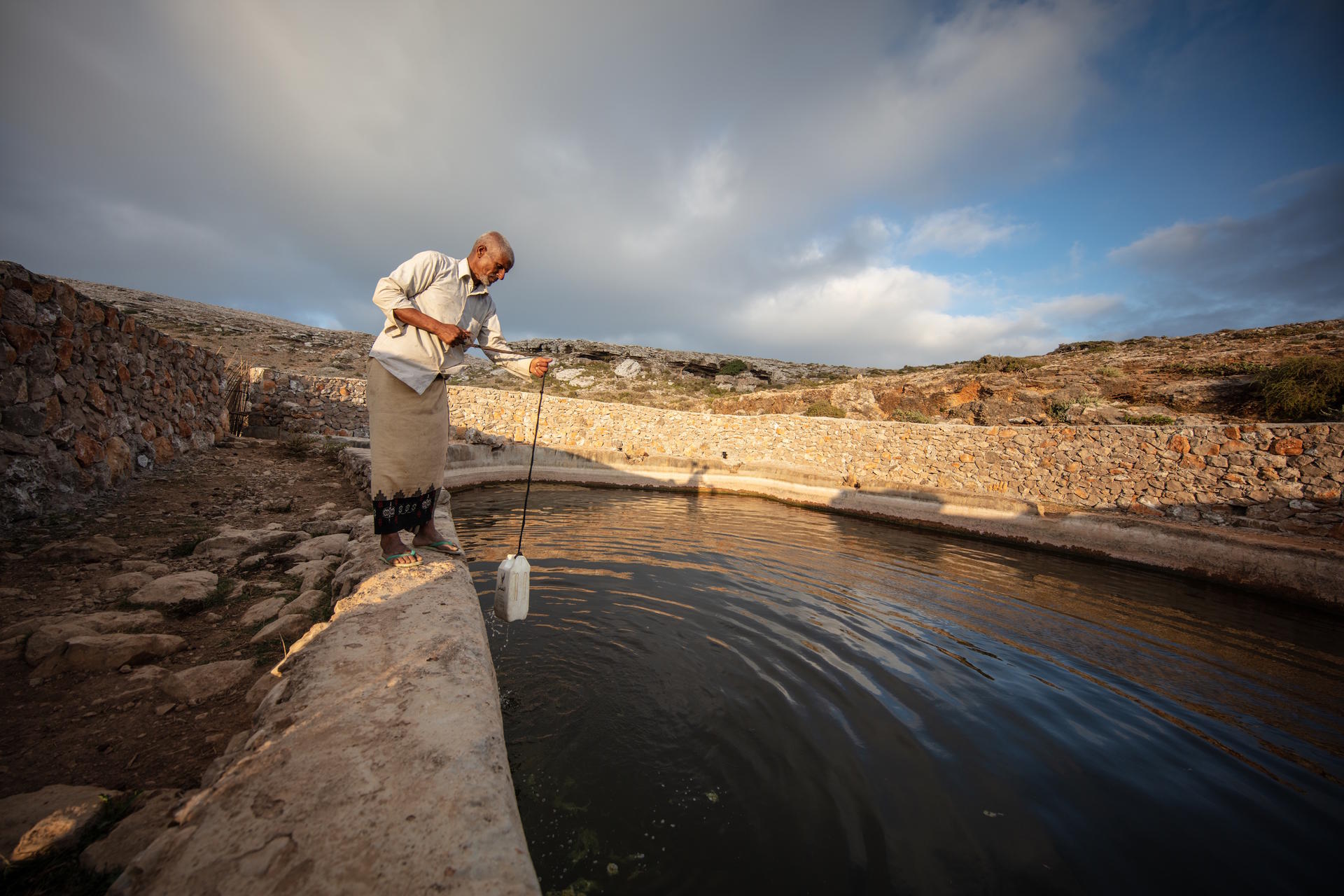 A person stands by a stone-lined water reservoir, testing the water with a device.