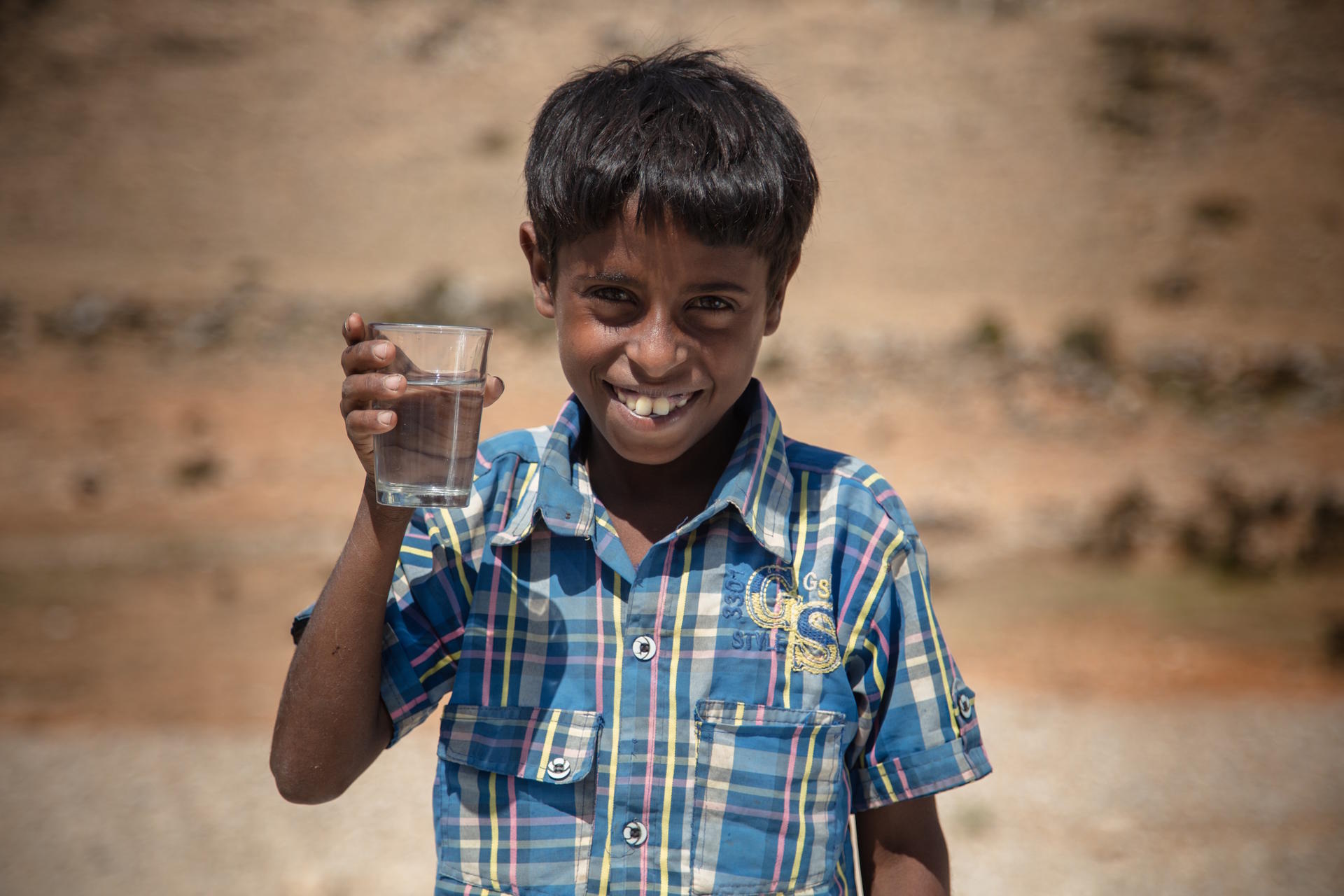 A smiling boy in a checked shirt holds up a glass of water outdoors.
