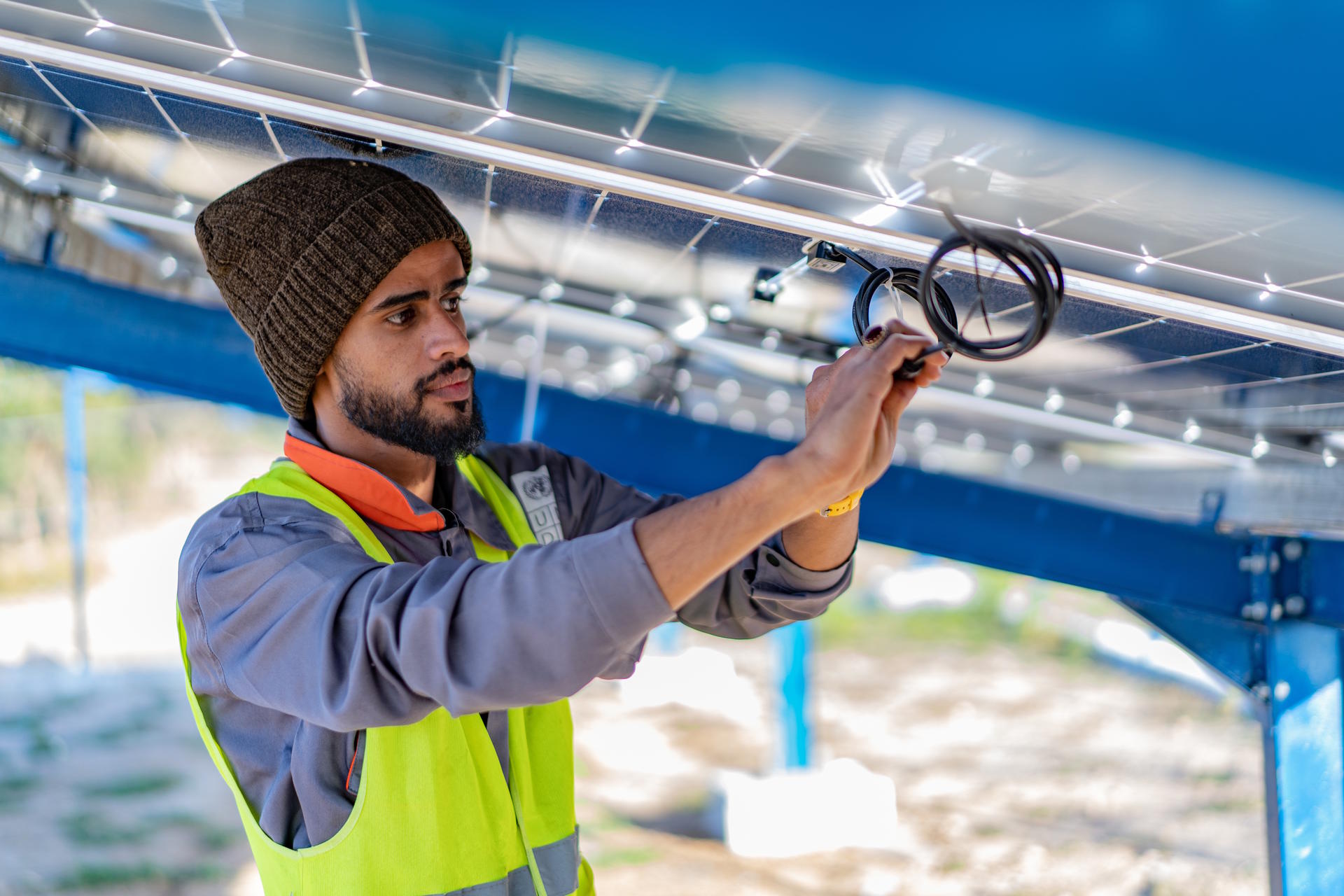 Man wearing a beanie and safety vest working on solar panel wiring outdoors.