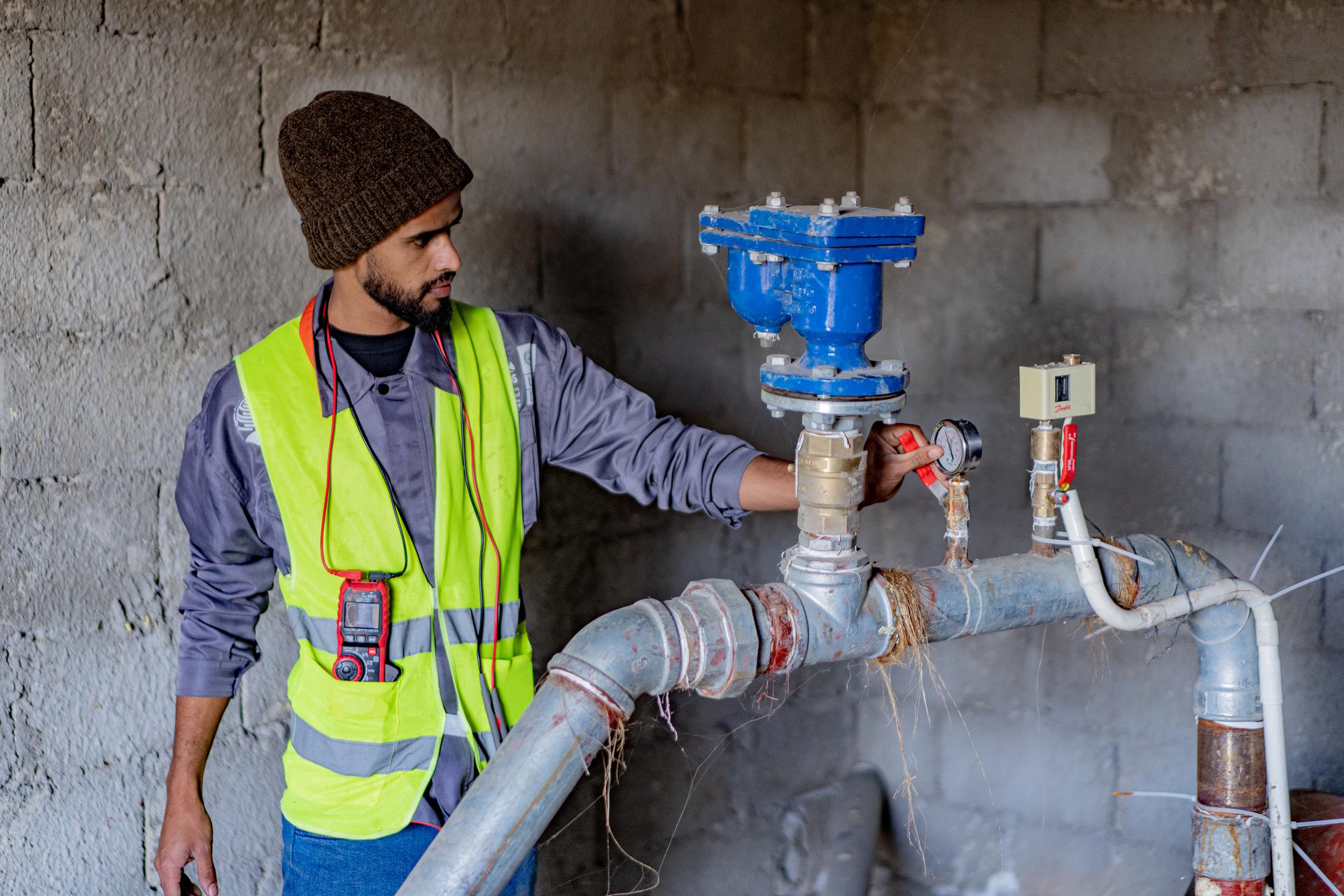 A worker in a yellow safety vest and beanie inspects a blue valve in a construction site.