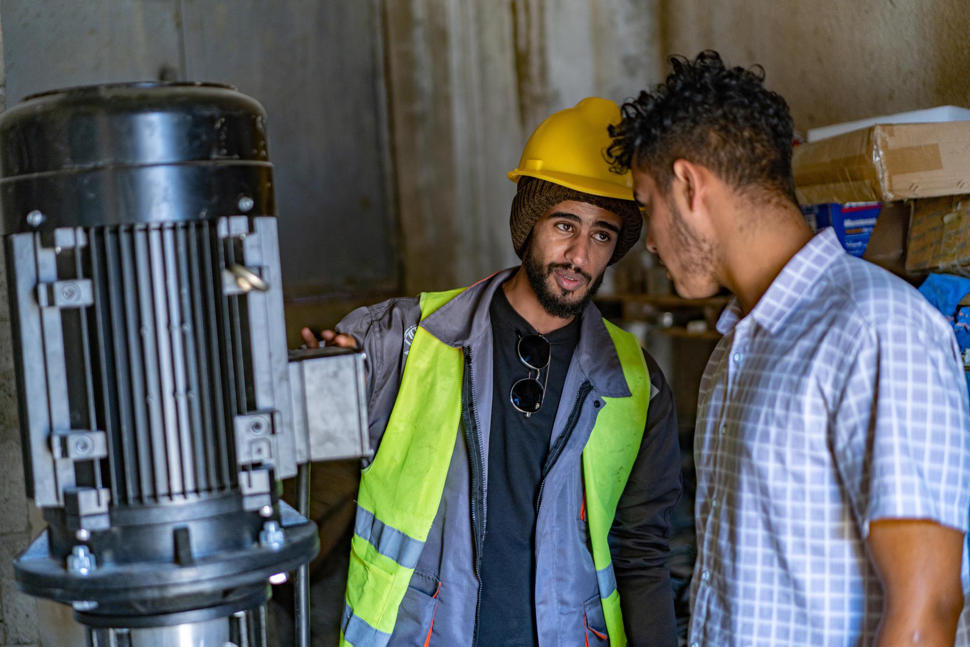 Two men discussing equipment in a workshop; one wearing a hard hat and safety vest.