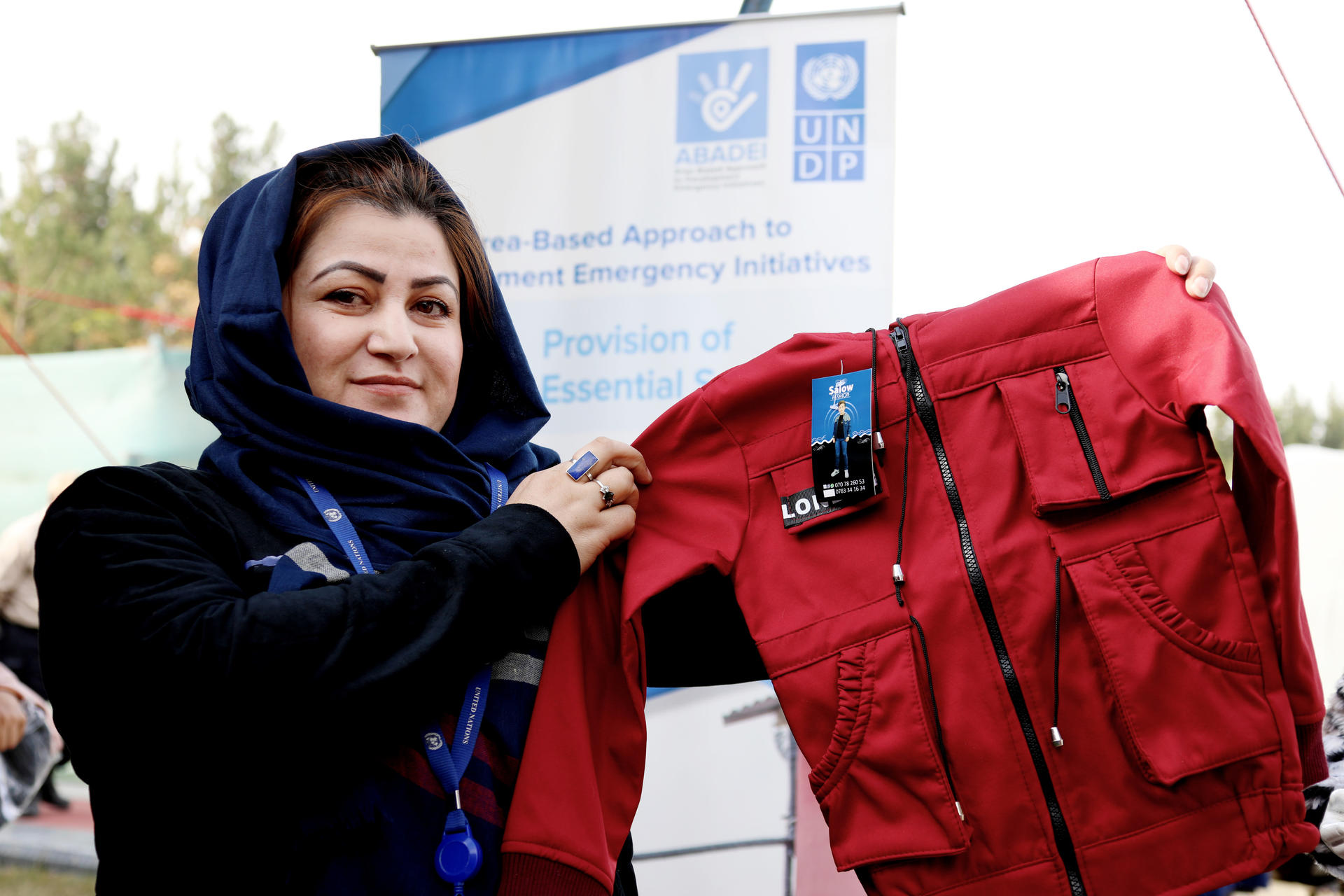 A woman in a headscarf holds up a red jacket, smiling, with a banner in the background.