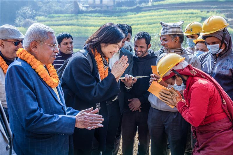 A group of people, some in hard hats, greet and interact with each other outdoors, with greenery in the background.
