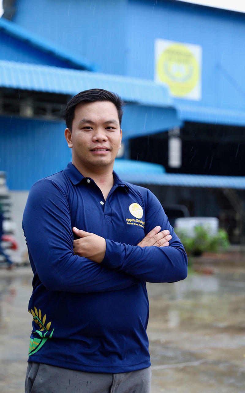 A man with folded arms stands confidently in front of a blue building on a rainy day.