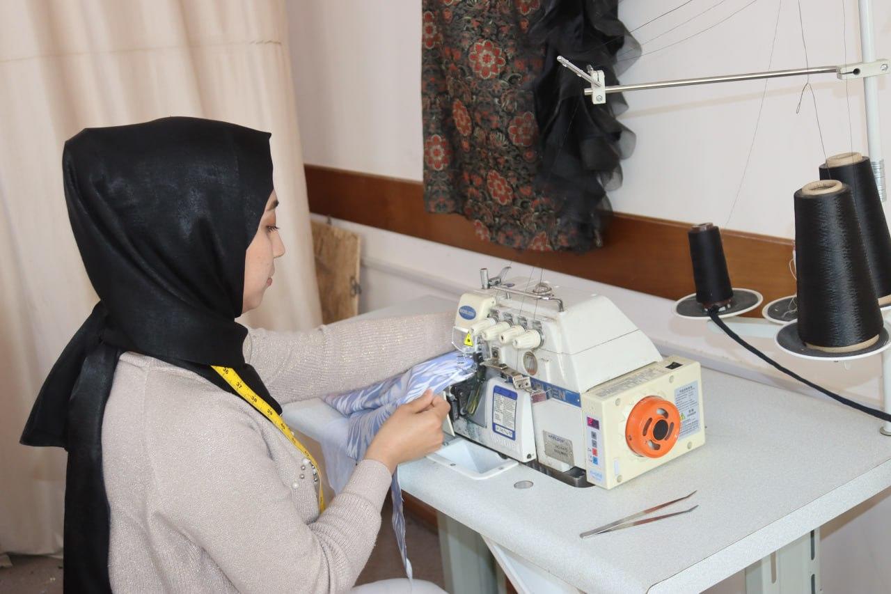 A woman in a hijab operates a sewing machine, focused on her work in a bright room.