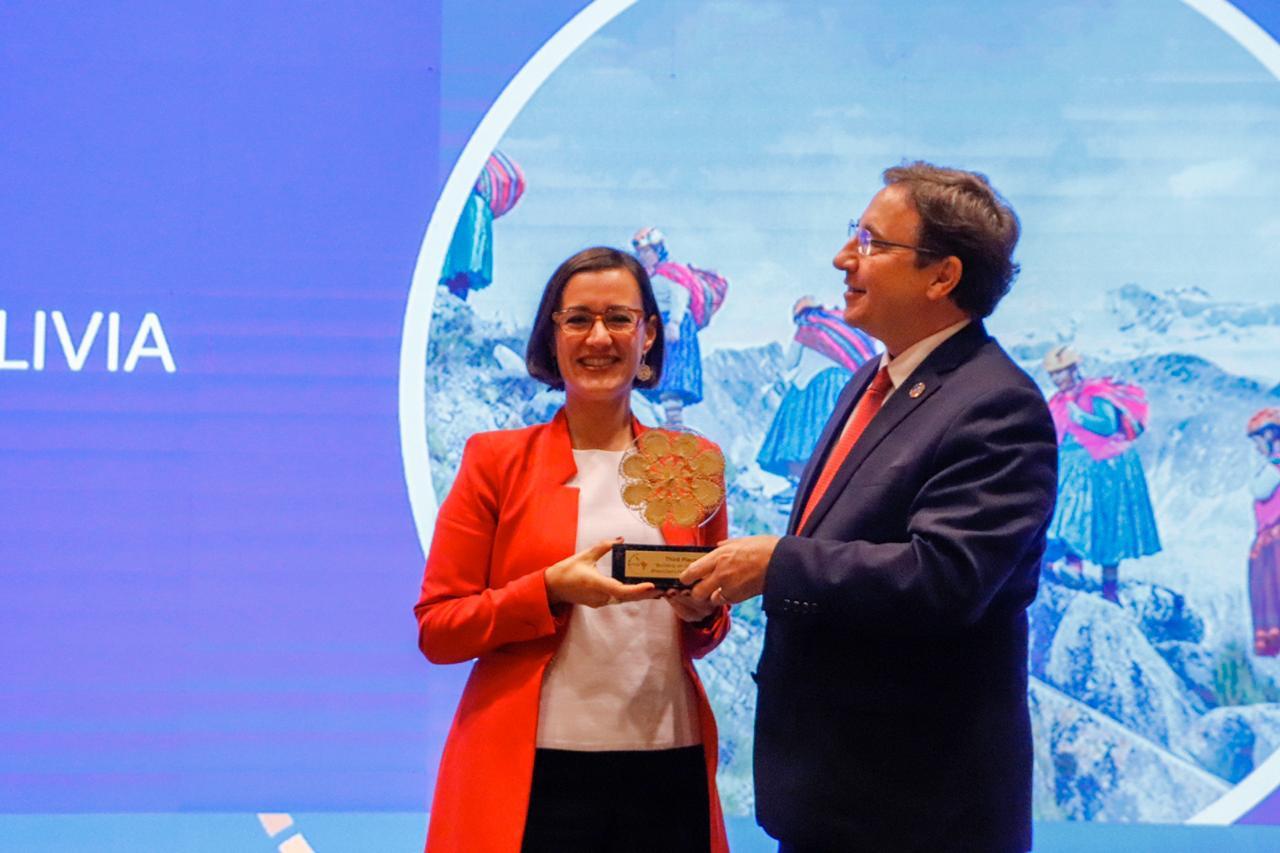 Two individuals are smiling and exchanging a trophy on a stage with a colorful background.