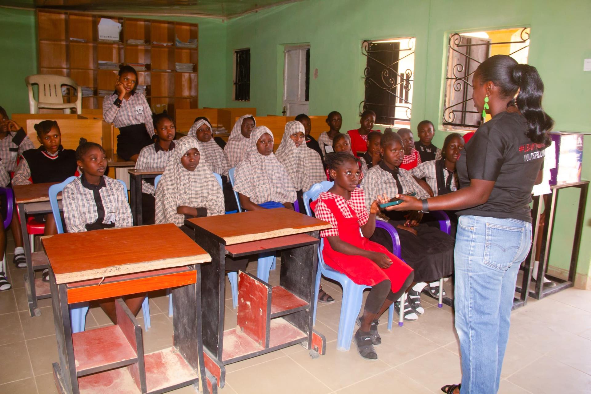 A group of students in uniforms listens attentively to a speaker in a classroom setting.