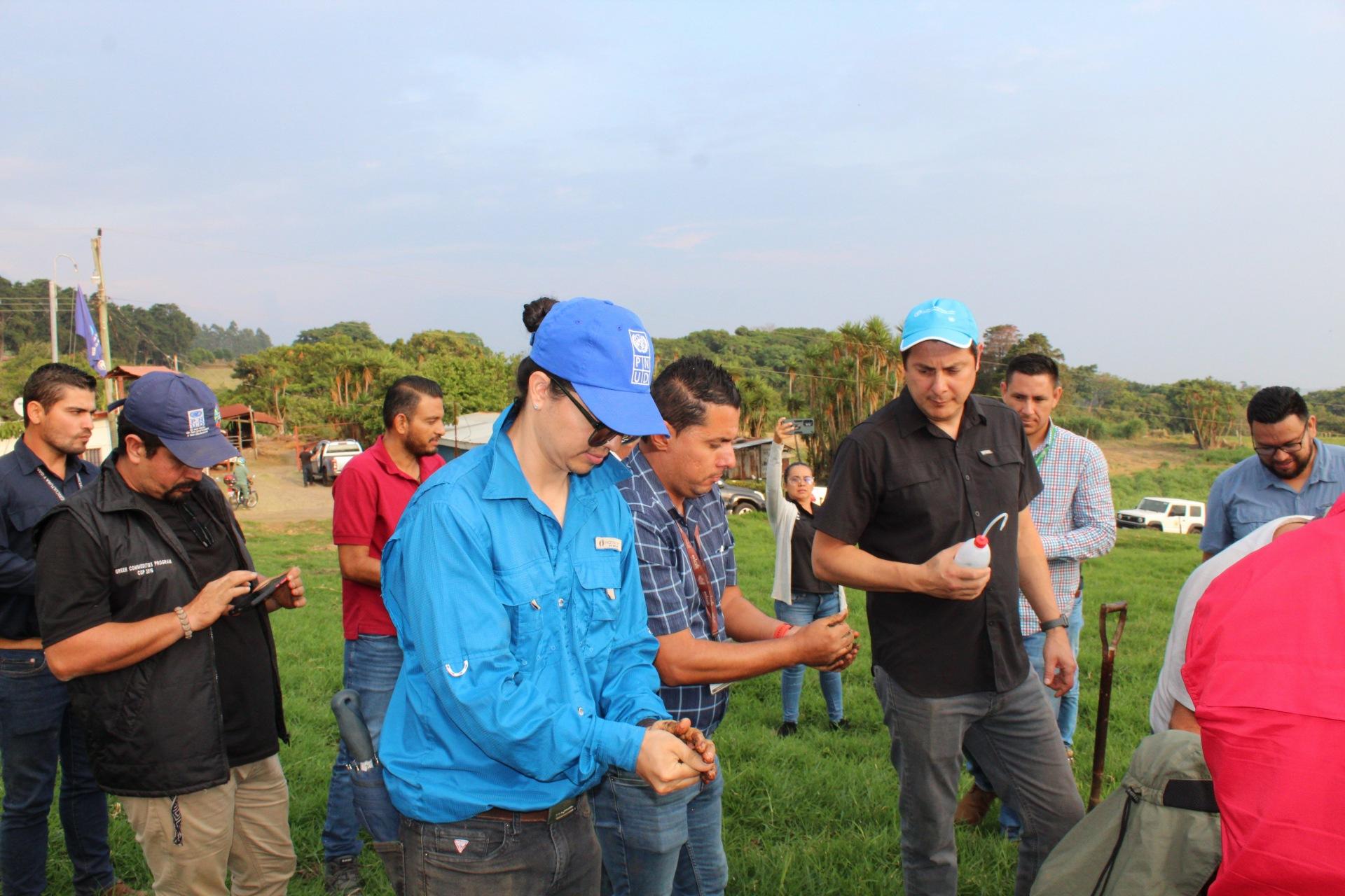 Group of people in a field, discussing and handling materials, with greenery in the background.