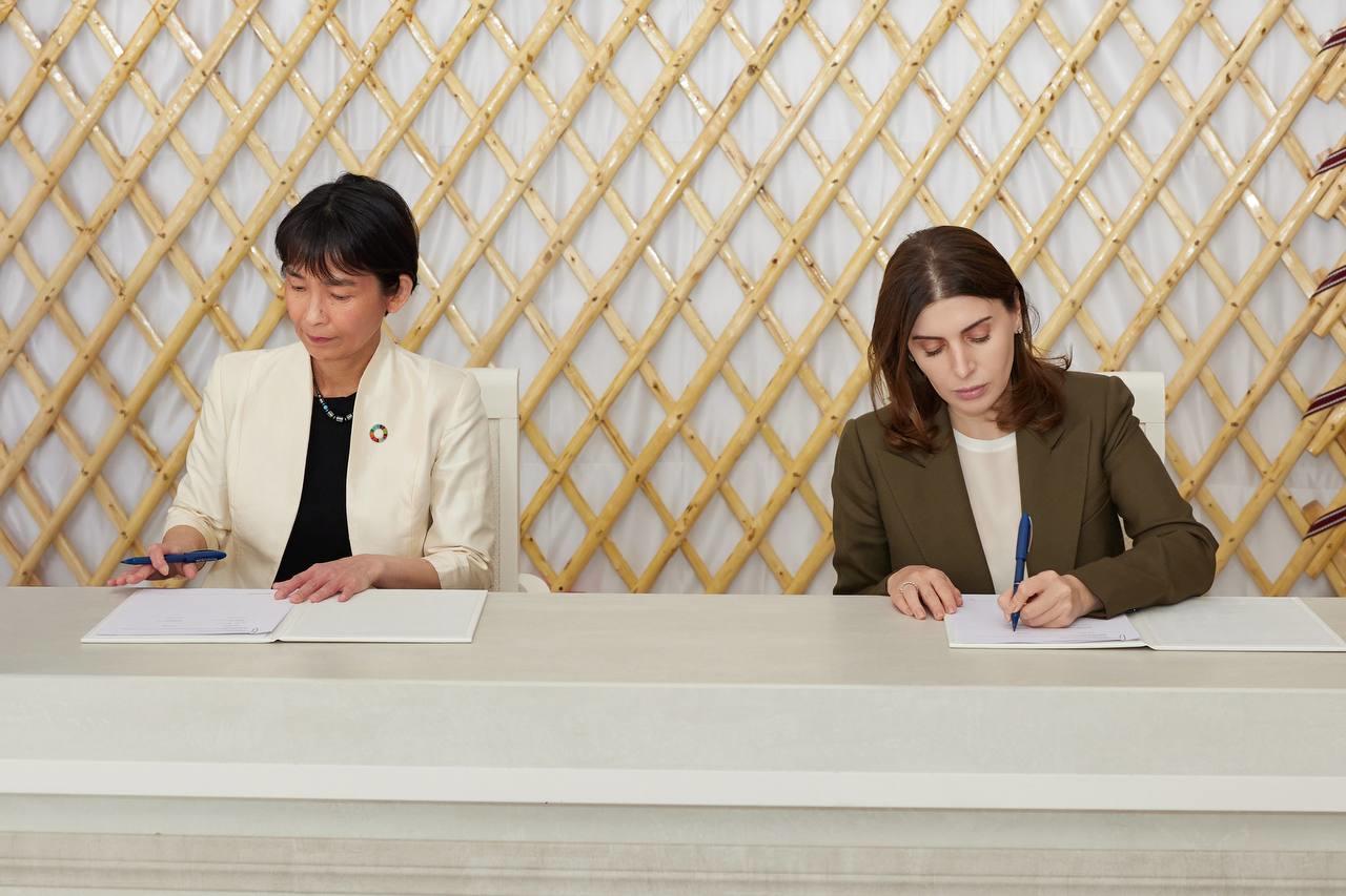 Two women signing documents at a table, with a geometric gold-patterned background.