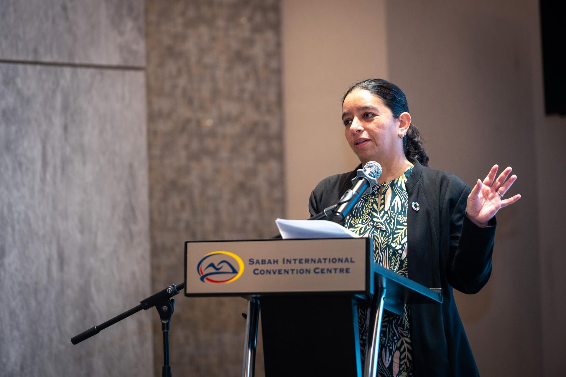 A speaker gesturing while presenting at a podium in a conference setting.