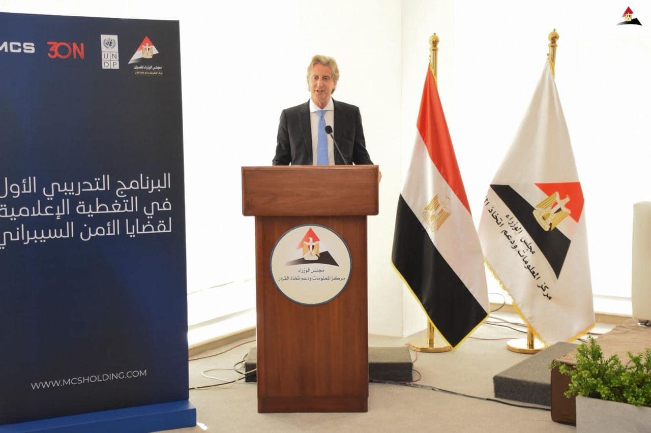 Man speaking at a podium with Egyptian flags and a blue backdrop displaying text.