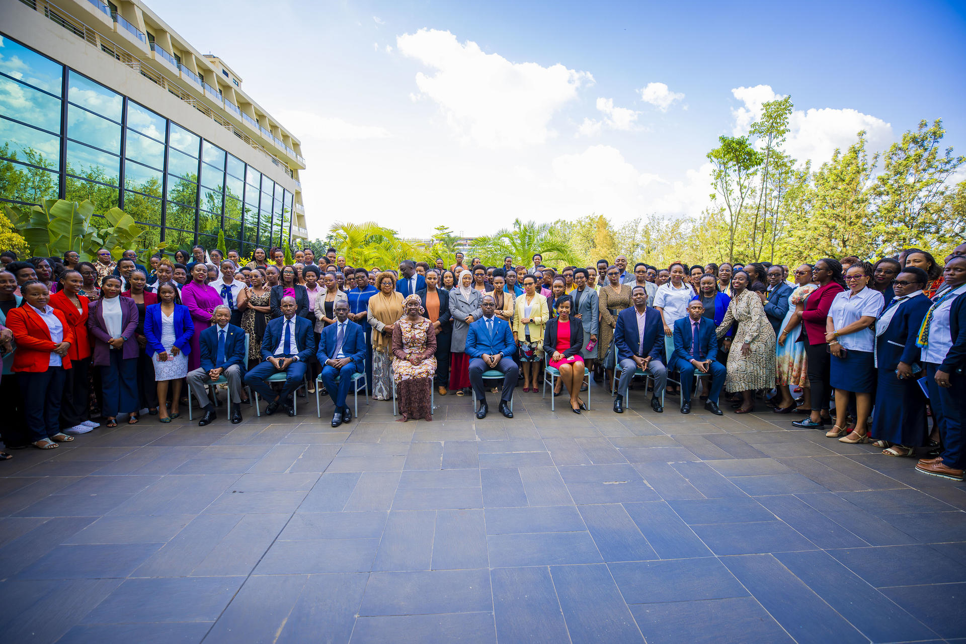 A large group of people gathered outdoors, smiling, with greenery and a modern building in the background.