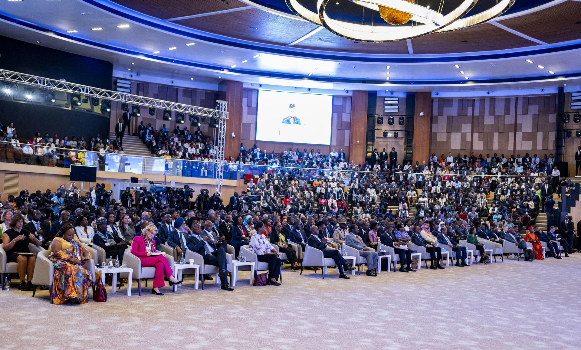 Crowd seated in a large conference hall, with a stage and screen at the front.