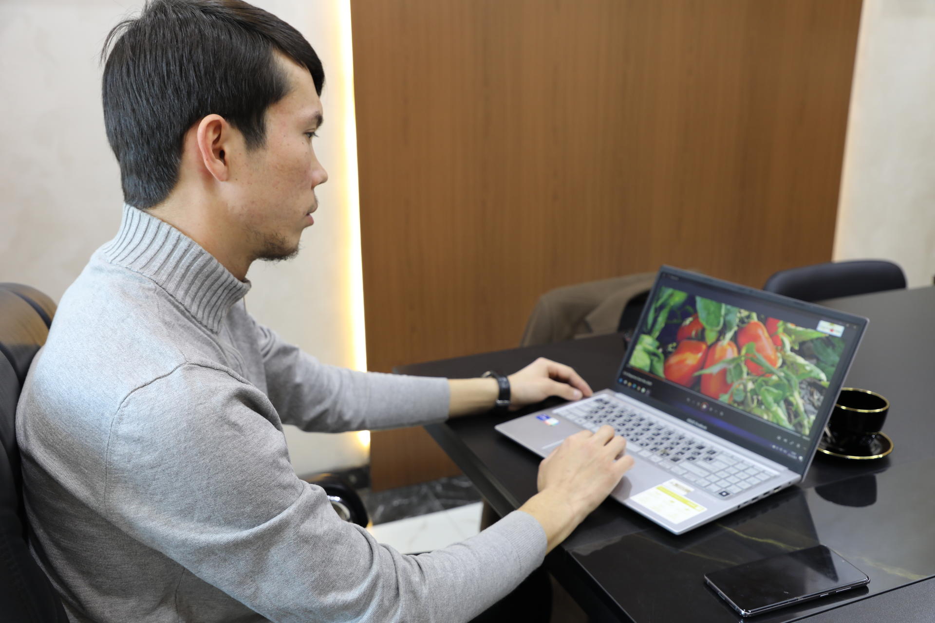 Man wearing a gray sweater works on a laptop at a desk with a coffee cup.