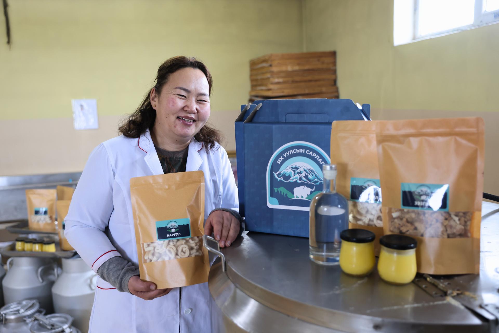 A woman in a lab coat smiles while holding a bag of products, surrounded by jars and packages.