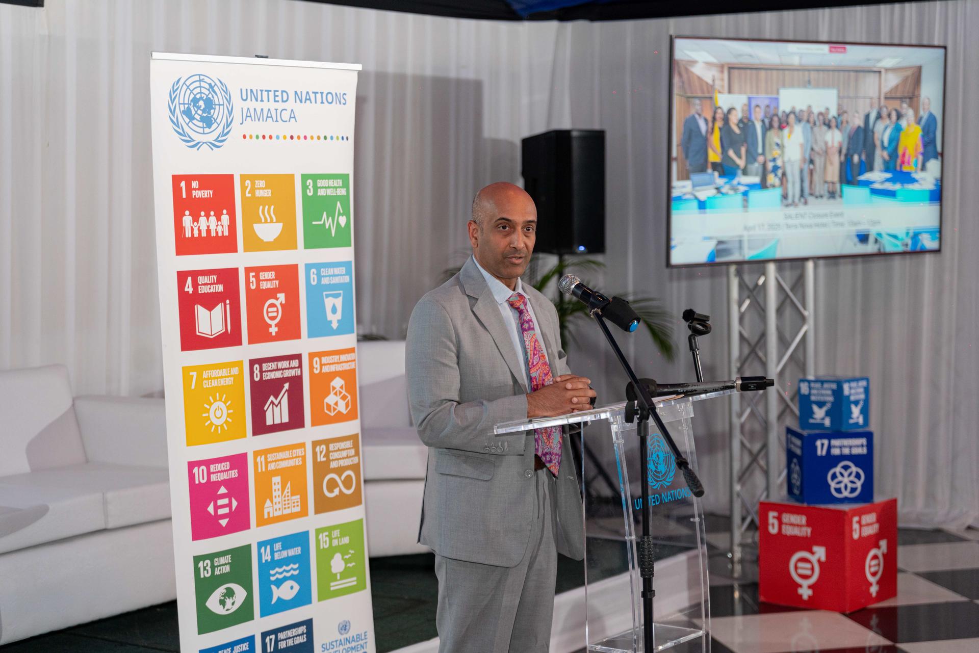 A man in a light gray suit speaks at a podium with UN branding and colorful goal icons behind him.